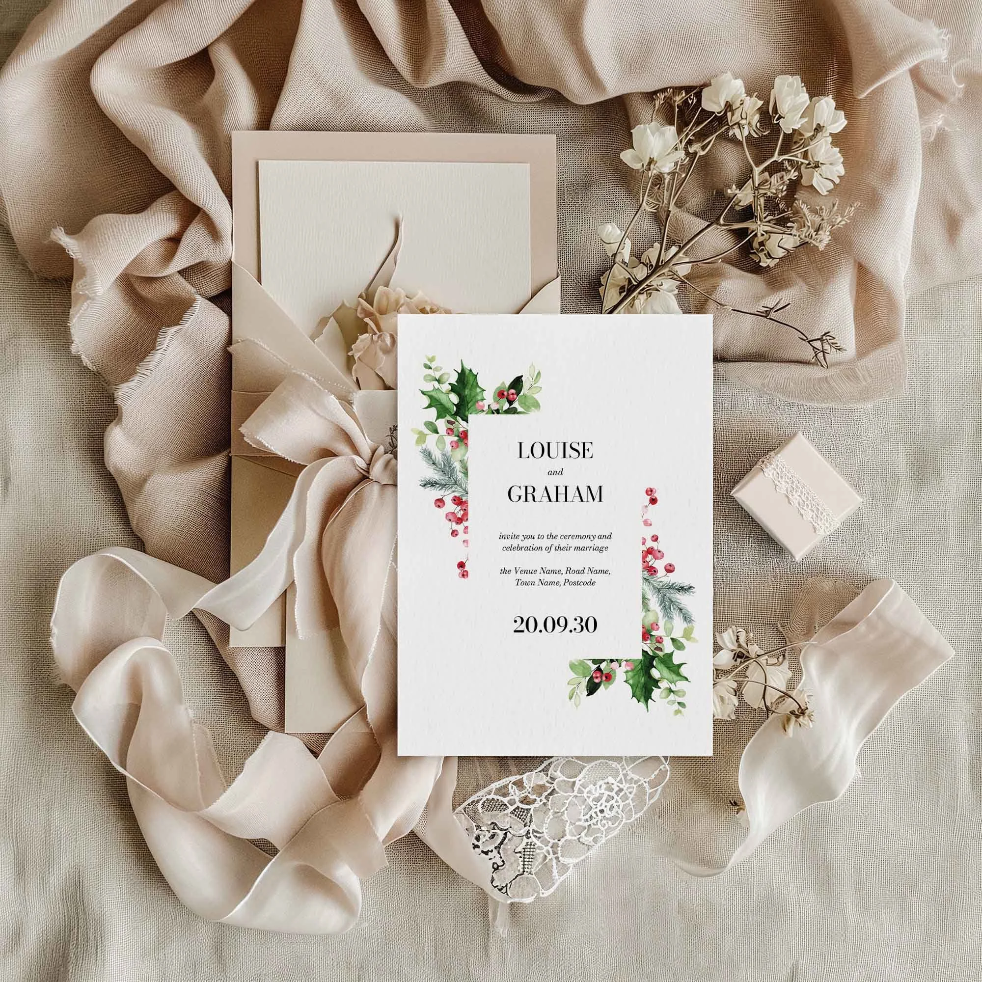 Festive Christmas wedding invitation flat lay featuring a holly, red berry and poinsettia corner arrangement, displayed on cream linen with dried flowers and a small gift box