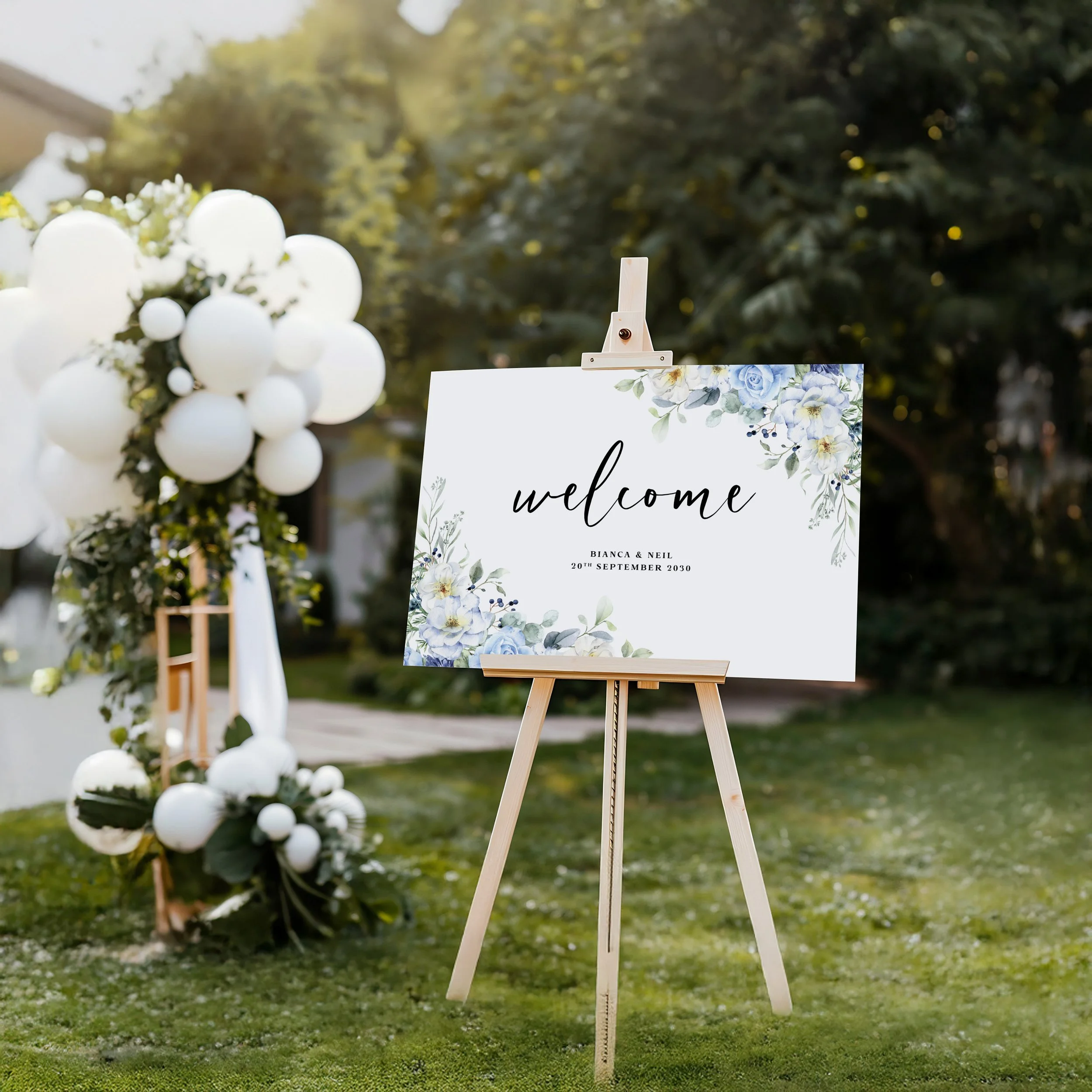 Wedding welcome sign with blue hydrangea illustration displayed on a wooden easel, surrounded by a white balloon arch and garden greenery