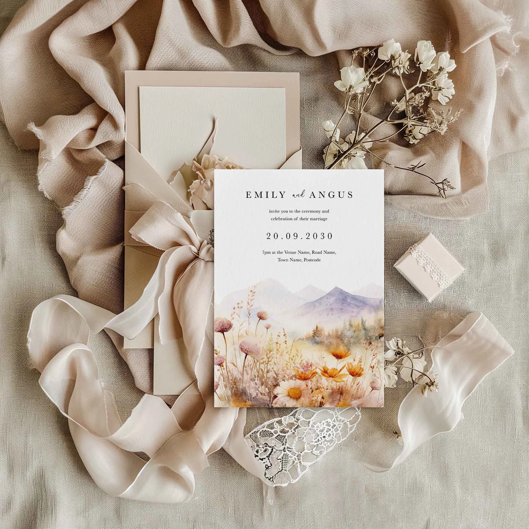 Scenic watercolour landscape wedding invitation flat lay featuring a purple mountain and golden wildflower meadow design, displayed on cream linen with dried flowers and a small gift box