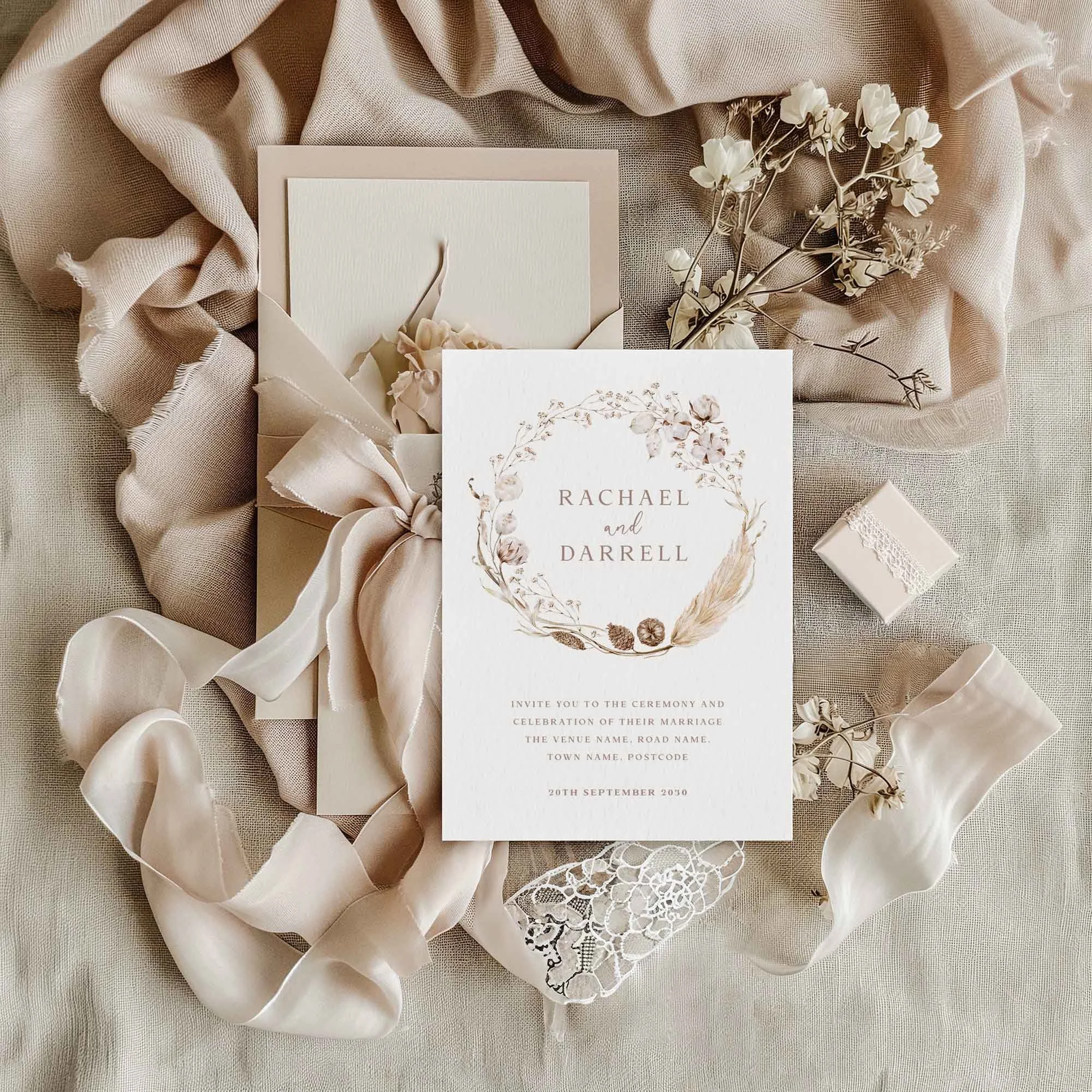 Boho dried flower wedding invitation flat lay featuring a neutral dried botanical wreath design, displayed on cream linen fabric with dried flowers and a small gift box
