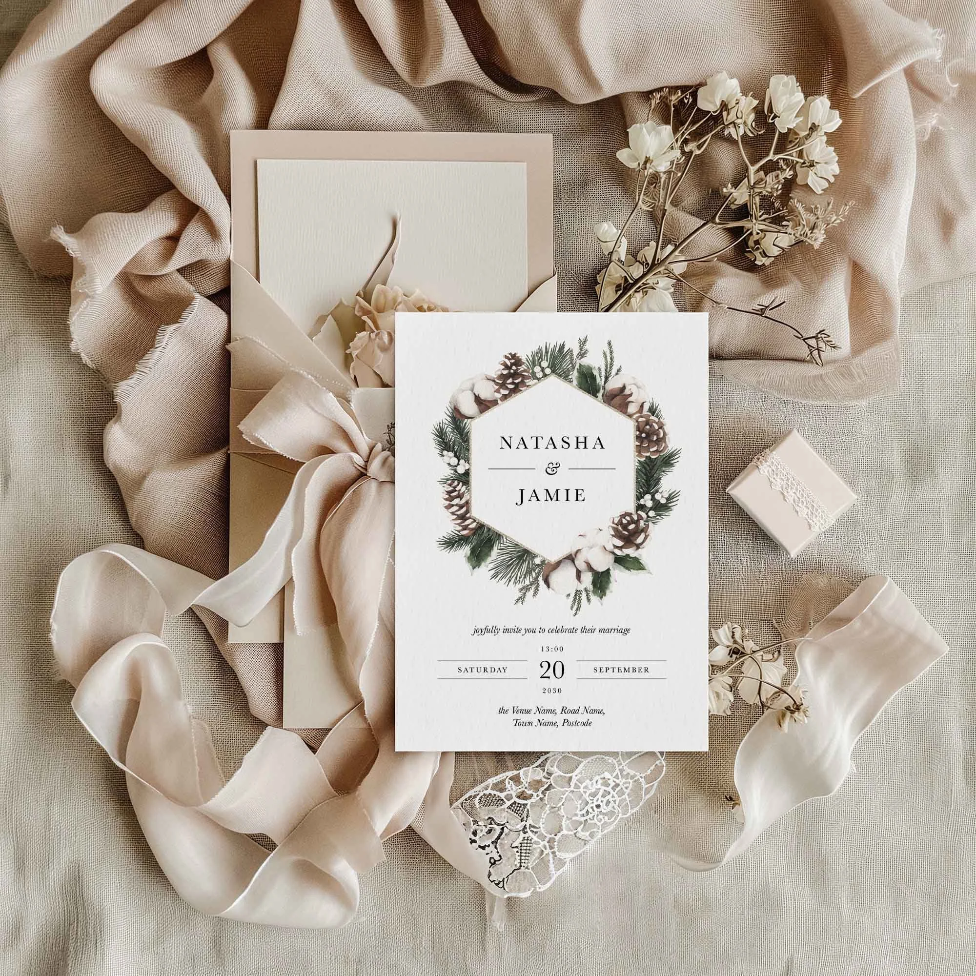 Woodland winter wedding invitation flat lay featuring a pine cone and cotton botanical design in a hexagonal frame, displayed on cream linen with dried flowers and a small gift box