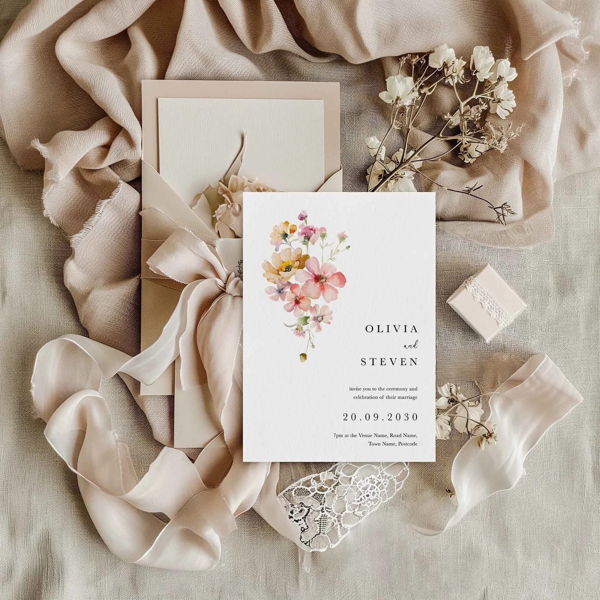 Blush pink watercolour wildflowers wedding invitation flat lay, displayed on cream linen fabric with dried flowers and a small gift box