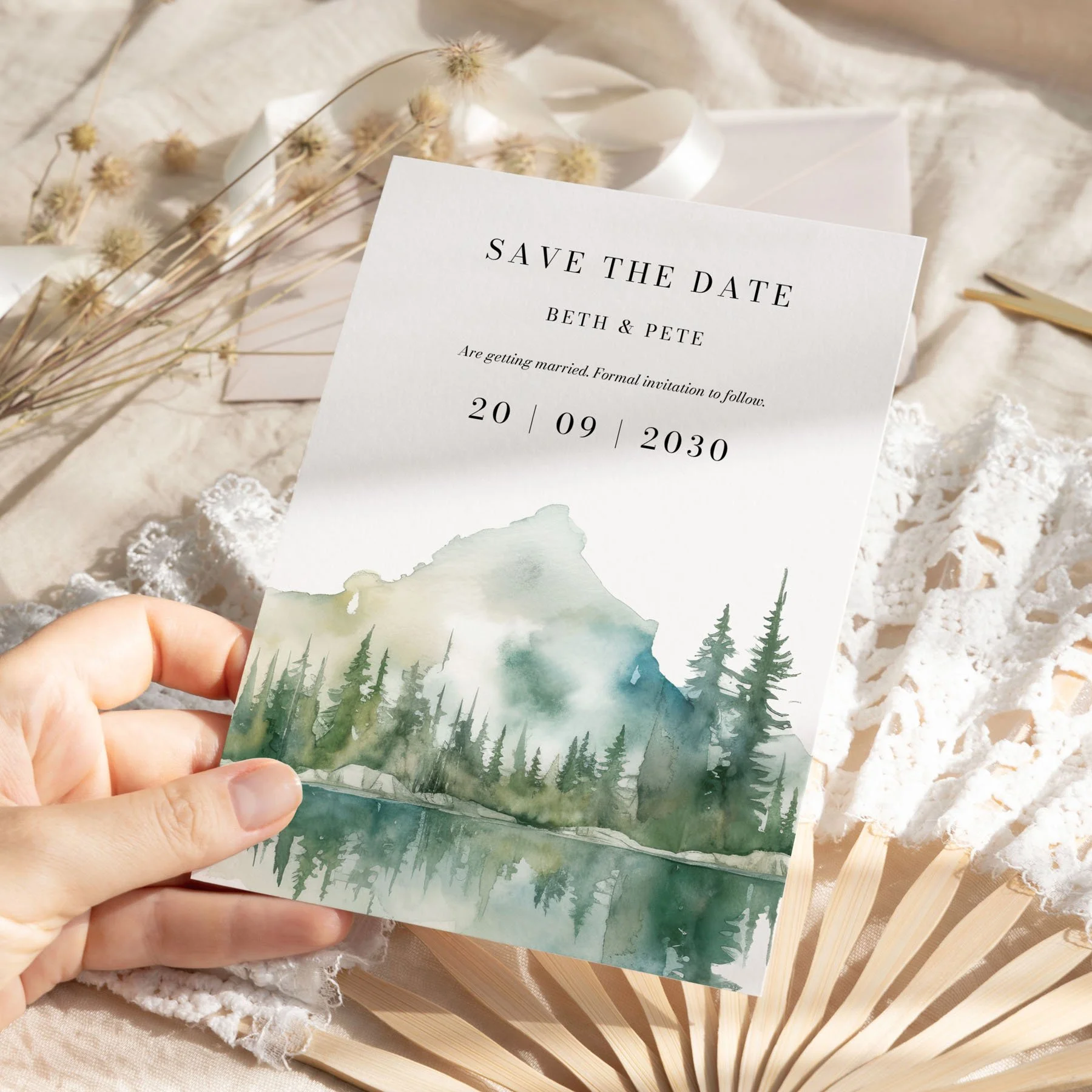 Hand holding Wilderness save the date card with watercolour mountain lake design