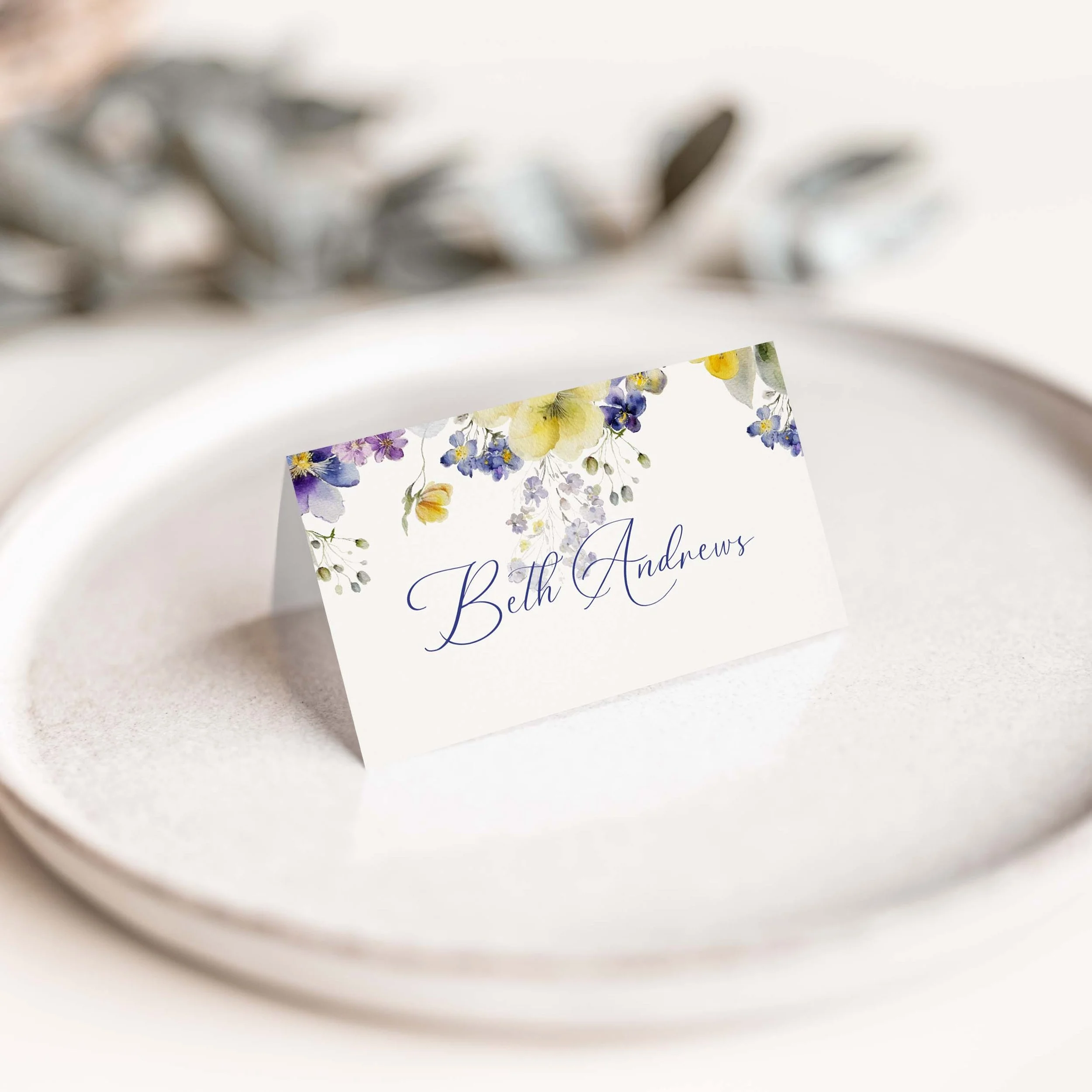 Violet wedding place card with purple violet and yellow botanical floral arrangement and flowing script guest name on luxury off-white card