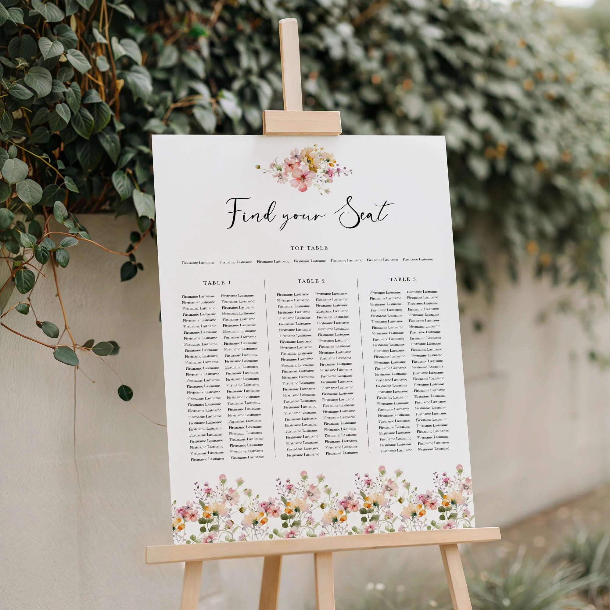 Bloom wedding seating plan displayed on a wooden easel, featuring a scattered pastel wildflower border with multiple guest tables