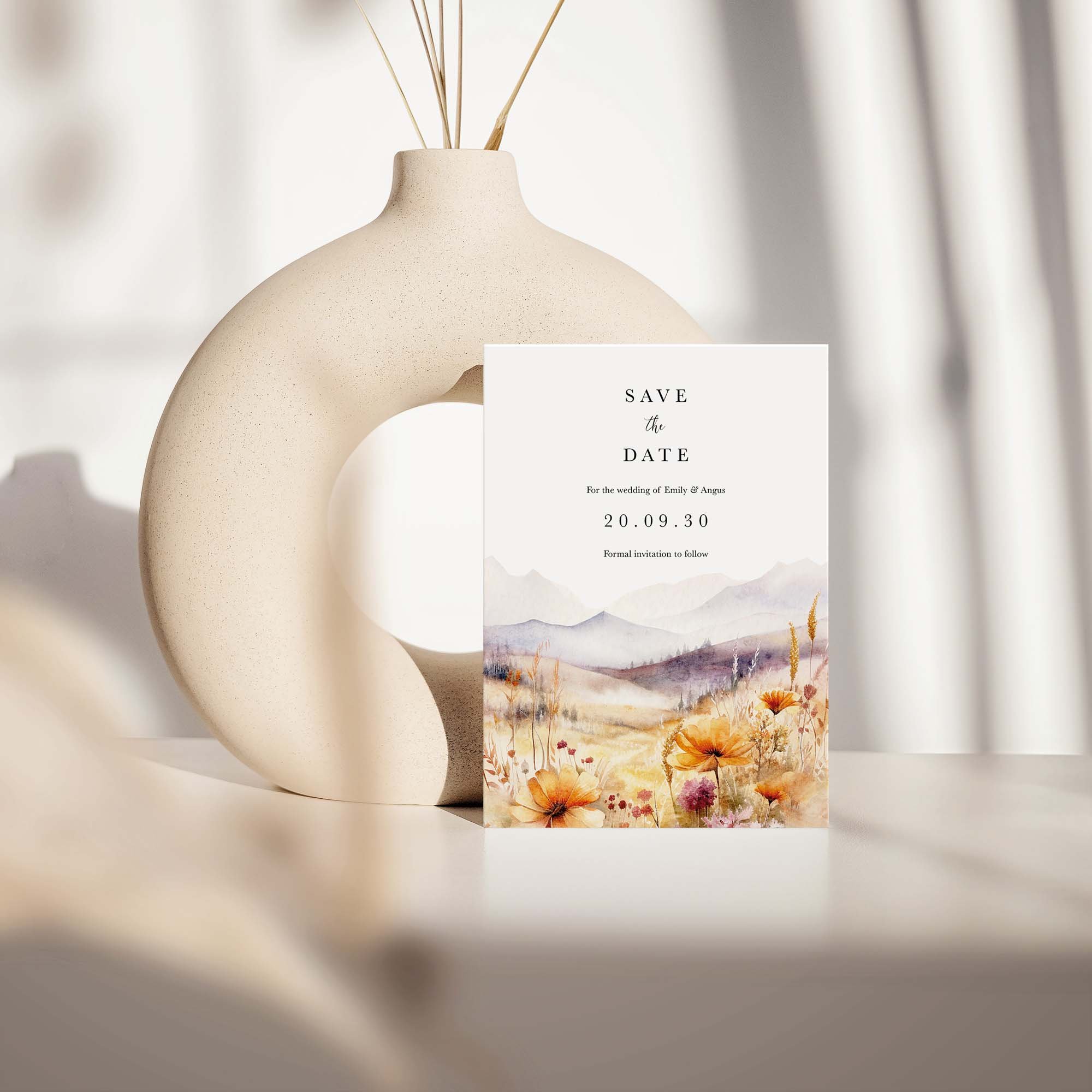 Scenic save the date card with watercolour landscape design featuring purple mountains and golden wildflower grasses, displayed against a cream ceramic vase on a neutral background