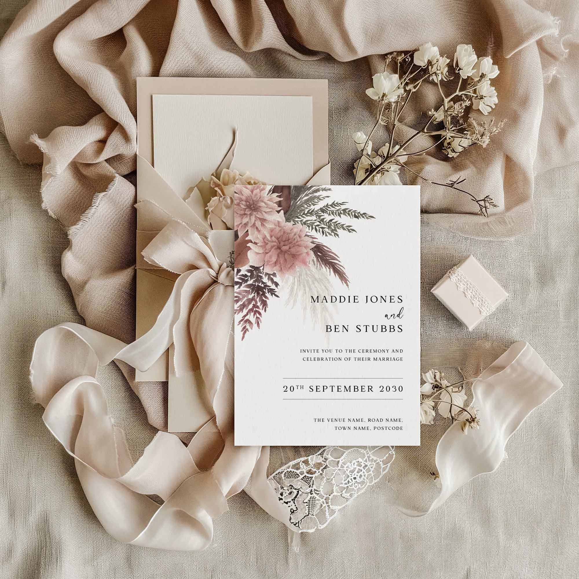 Blush Boho wedding invitation flat lay featuring pampas grass and blush pink dahlia corner arrangement, displayed on cream linen fabric with dried flowers and a small gift box