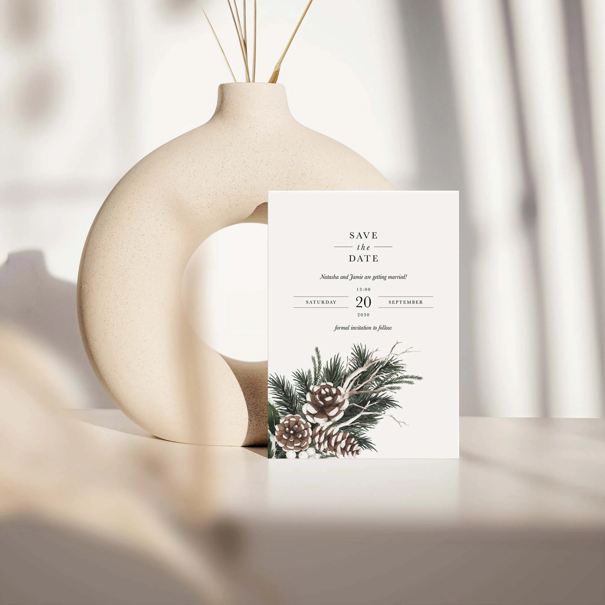 Woodland save the date card with pine cone and evergreen botanical design displayed against a cream ceramic vase on a neutral background