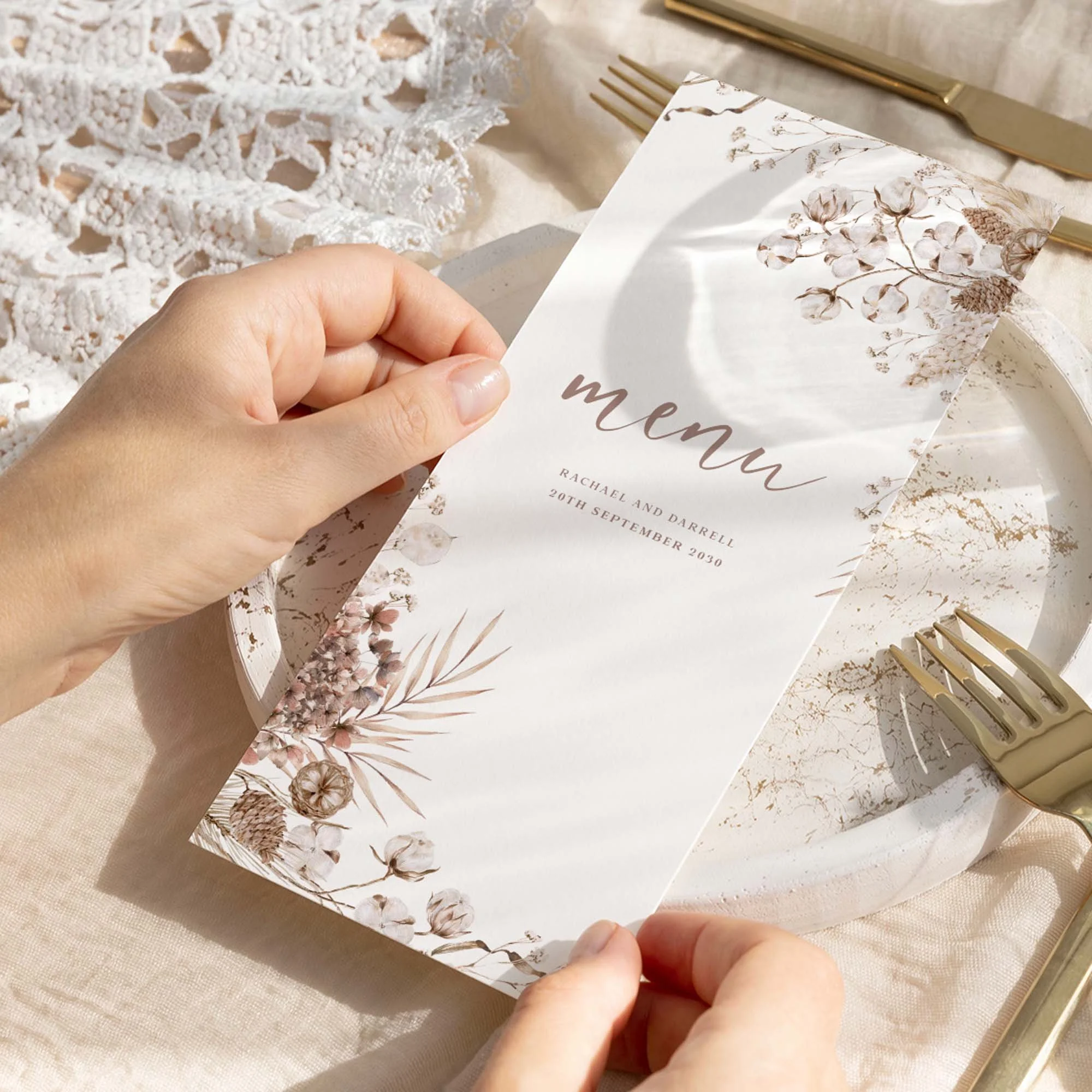 Hand holding a Boho wedding breakfast menu card with a dried botanical corner arrangement in warm ivory and taupe tones