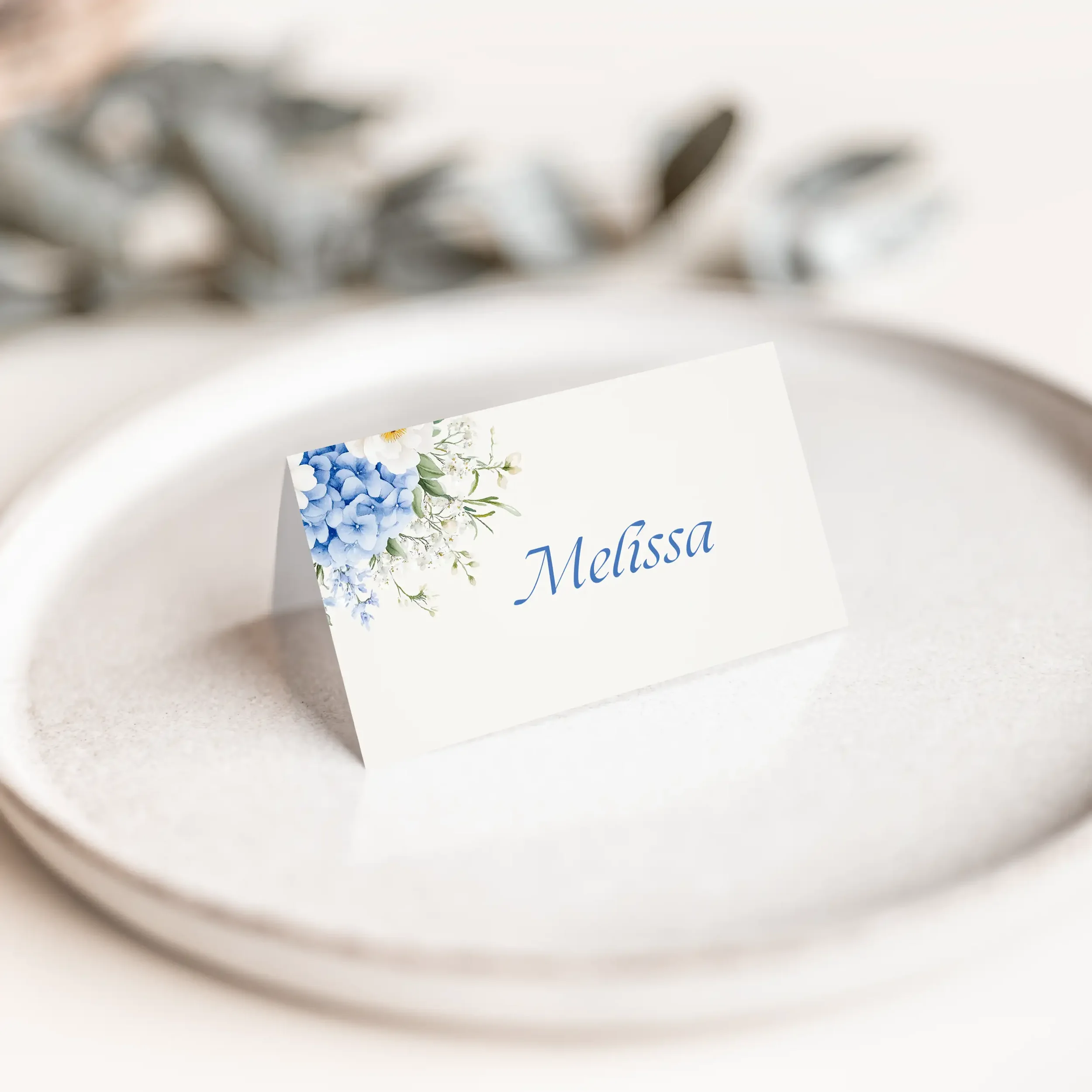 Serenity wedding place card with soft blue hydrangea floral illustration above flowing script guest name on luxury off-white card