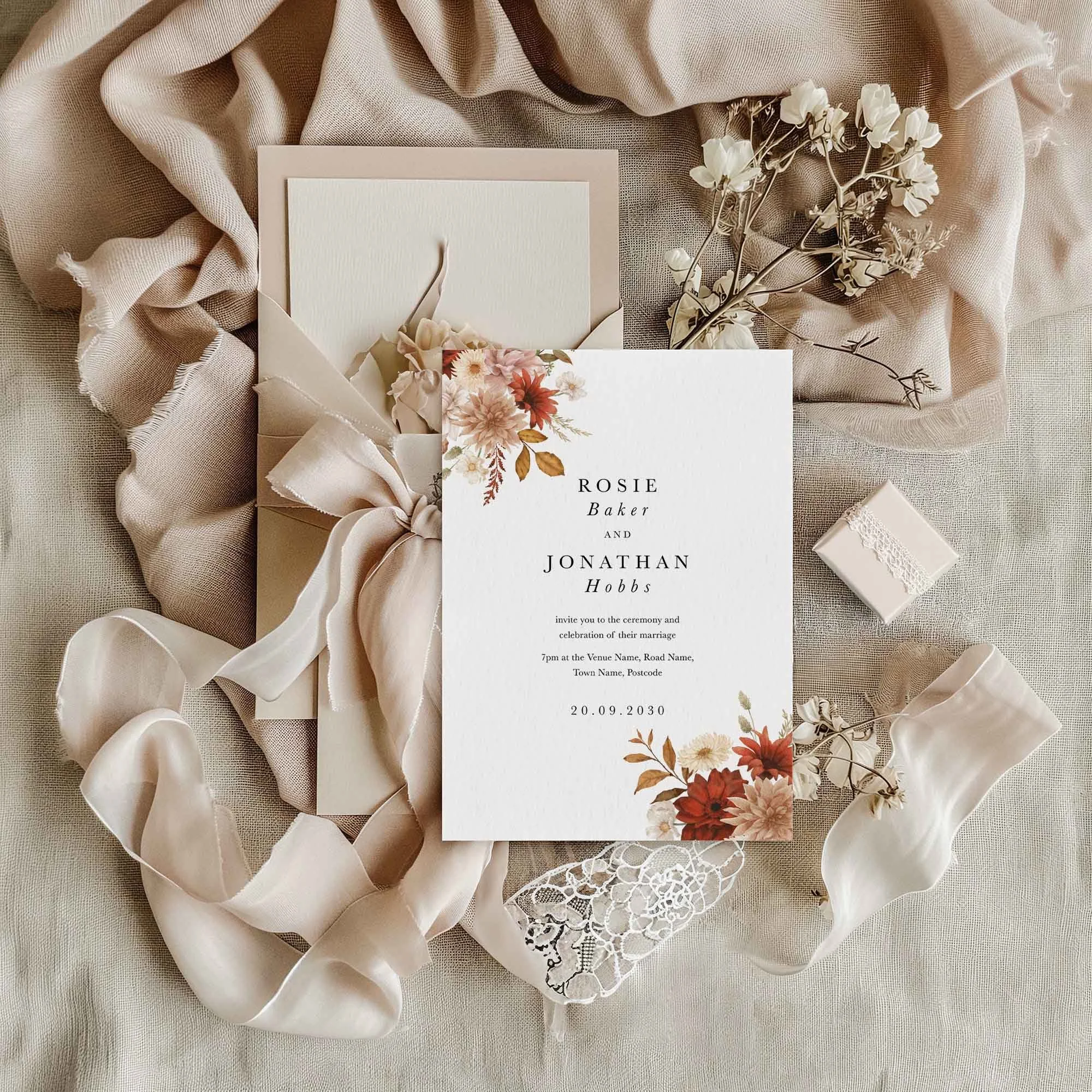 Rustic wedding invitation flat lay featuring a bold crimson dahlia and cream floral corner arrangement, displayed on cream linen with dried flowers and a small gift box
