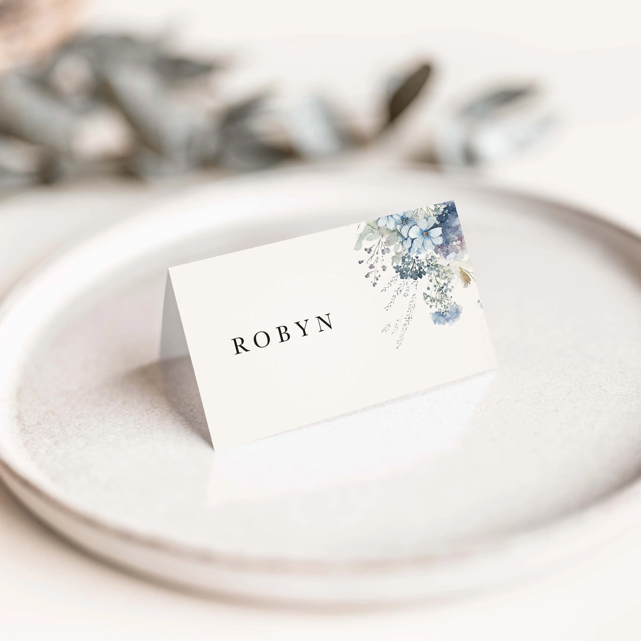 Dusty Blue wedding place card with soft blue floral botanical illustration and elegant serif typography on luxury off-white card