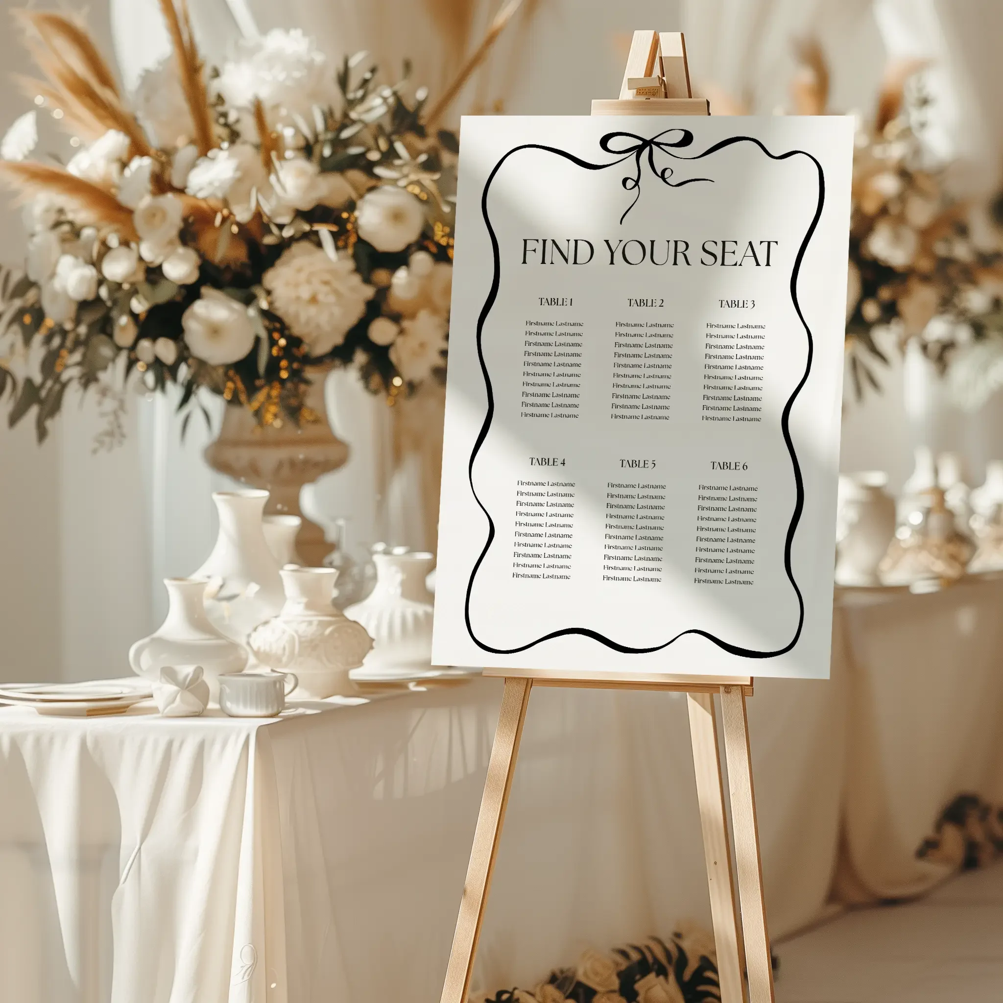 Wedding seating chart with decorative wavy border and bow design displayed on a wooden easel in a white floral wedding venue