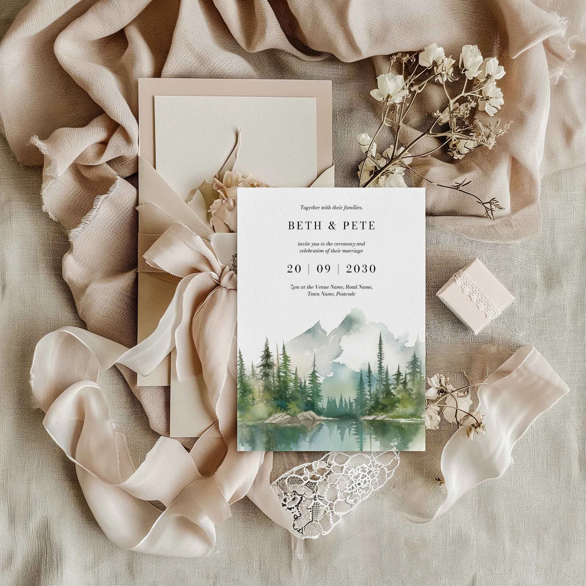 Wilderness wedding invitation flat lay featuring watercolour mountain lake and pine forest design, displayed on cream linen fabric with dried flowers and a small gift box