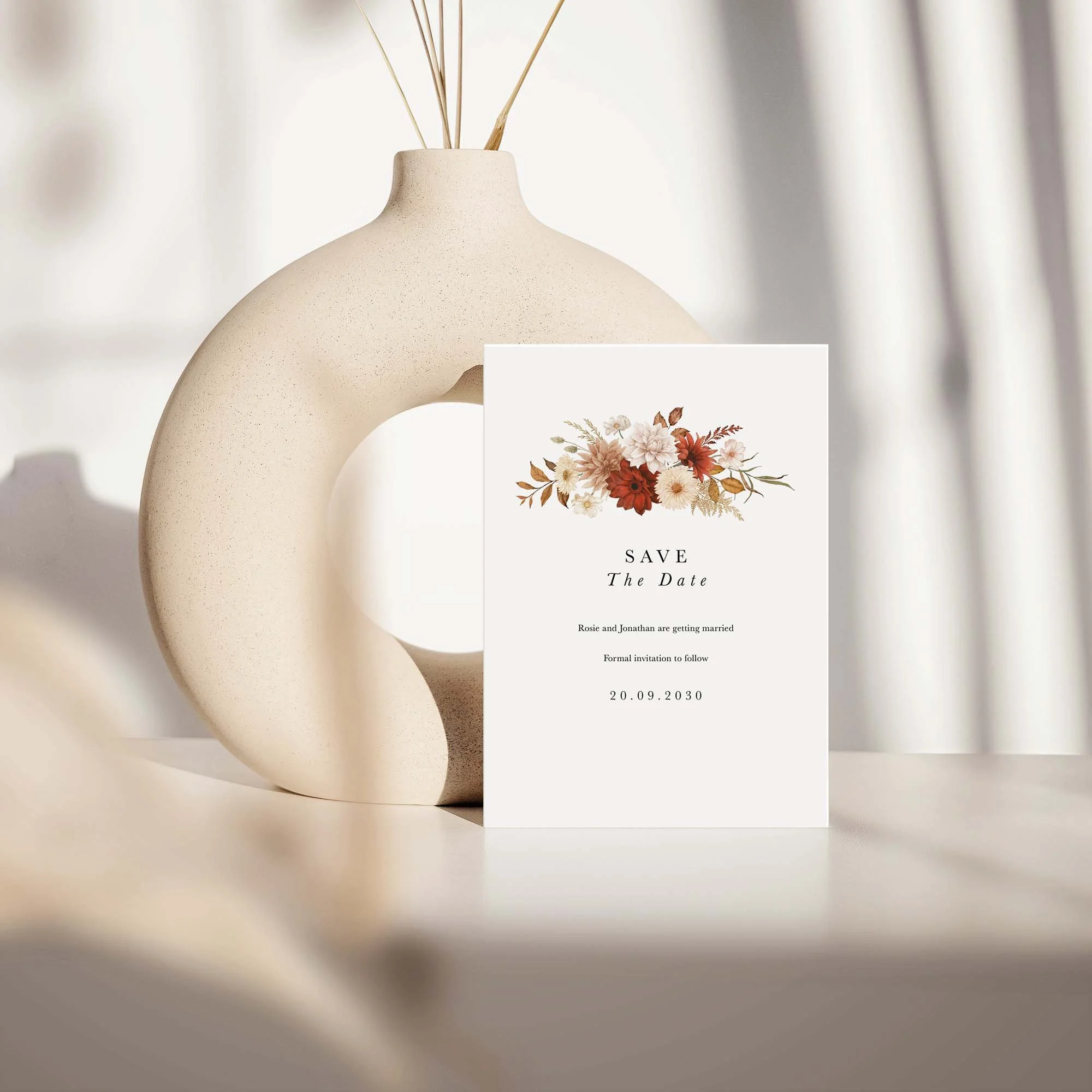 Rustic save the date card with crimson and cream floral design displayed against a cream ceramic vase on a neutral background