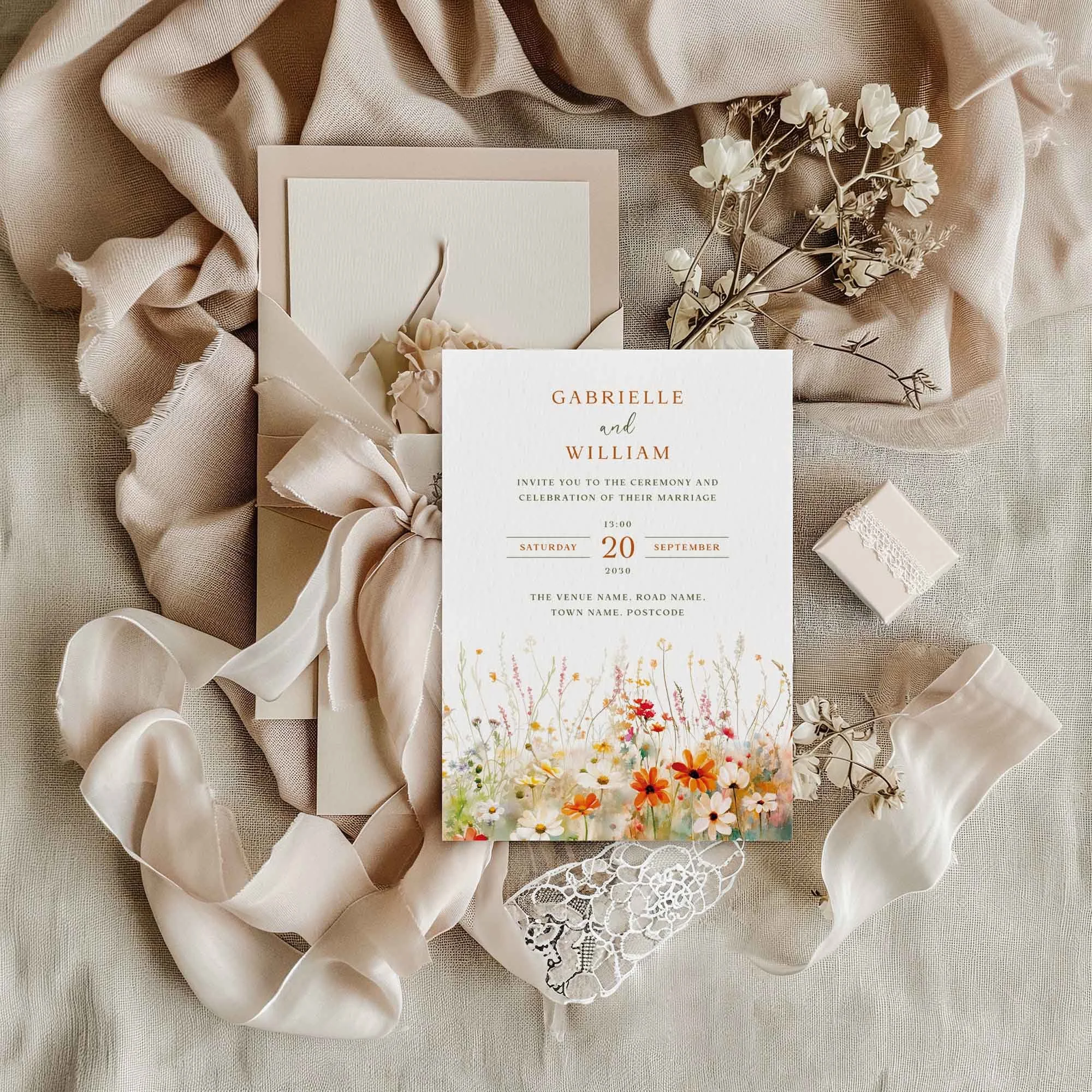 Field Vermilion wedding invitation flat lay featuring a vibrant orange and terracotta wildflower meadow border, displayed on cream linen with dried flowers and a small gift box