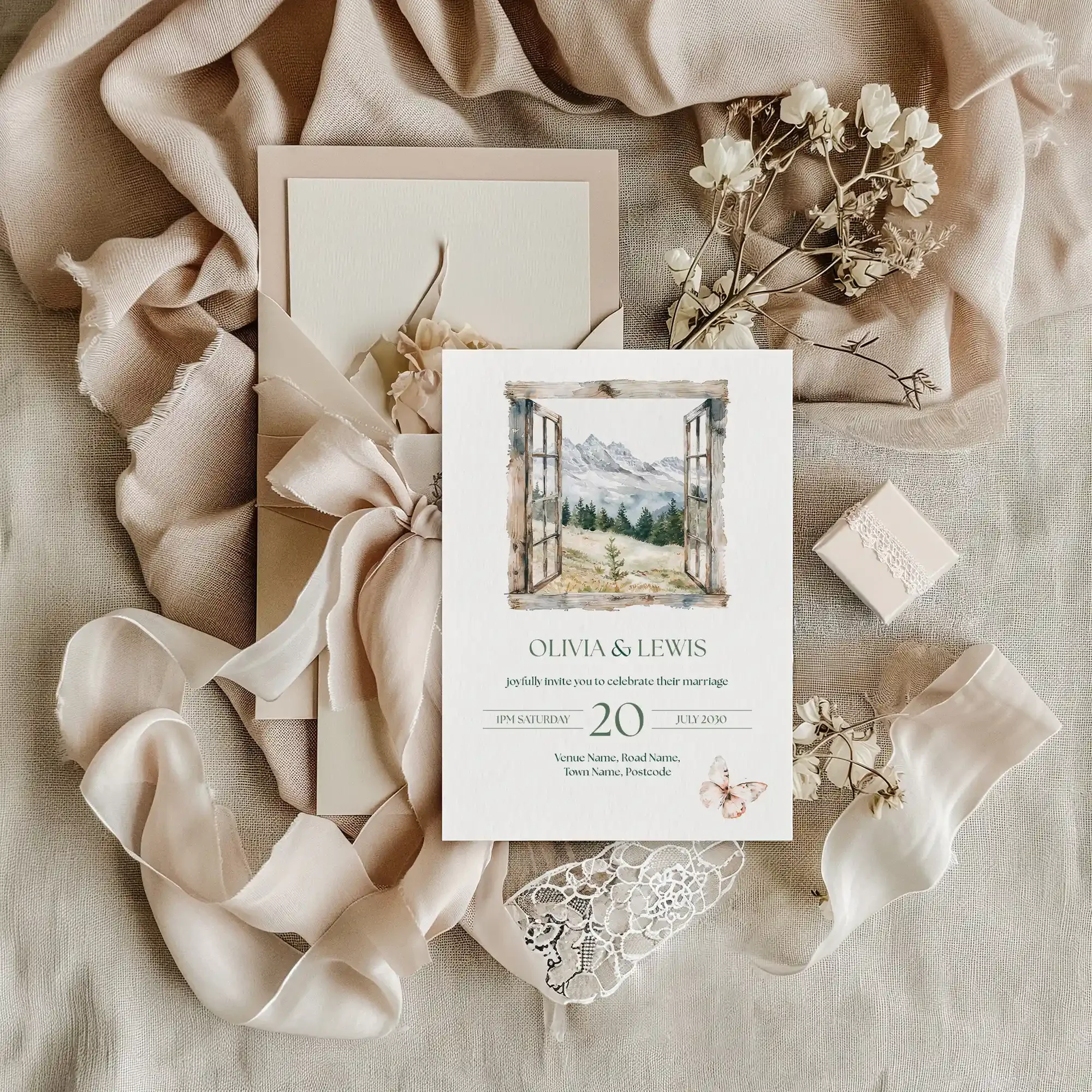 Adventure wedding invitation flat lay featuring illustrated mountain window design, displayed on cream linen fabric with dried flowers and a small gift box