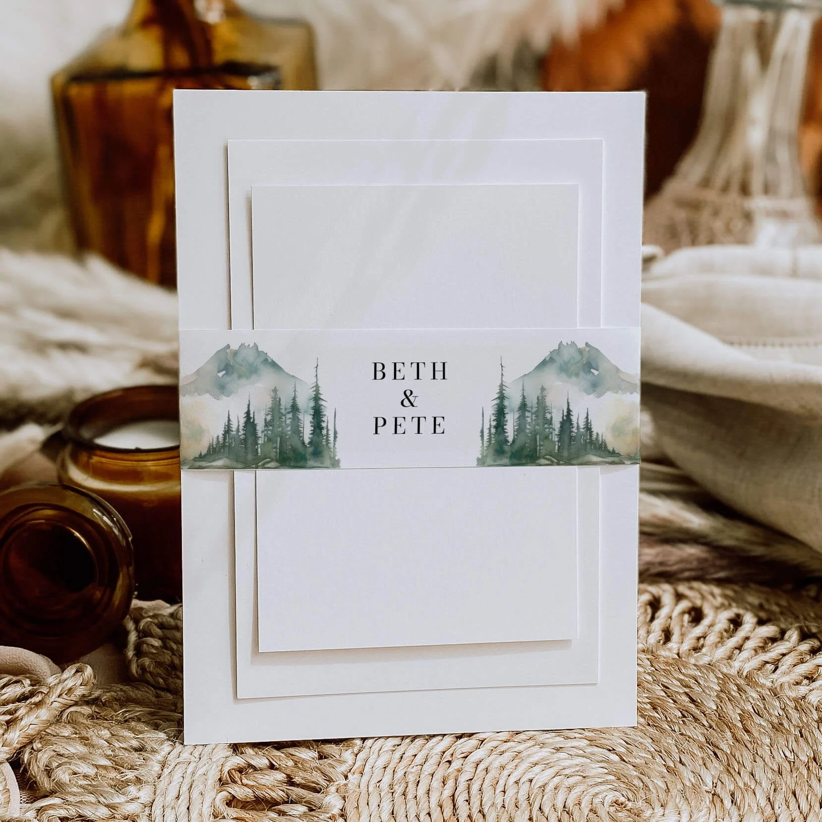 Wilderness personalised wedding belly bands with watercolour mountain and pine forest landscape illustration and serif names wrapped around wedding invitations