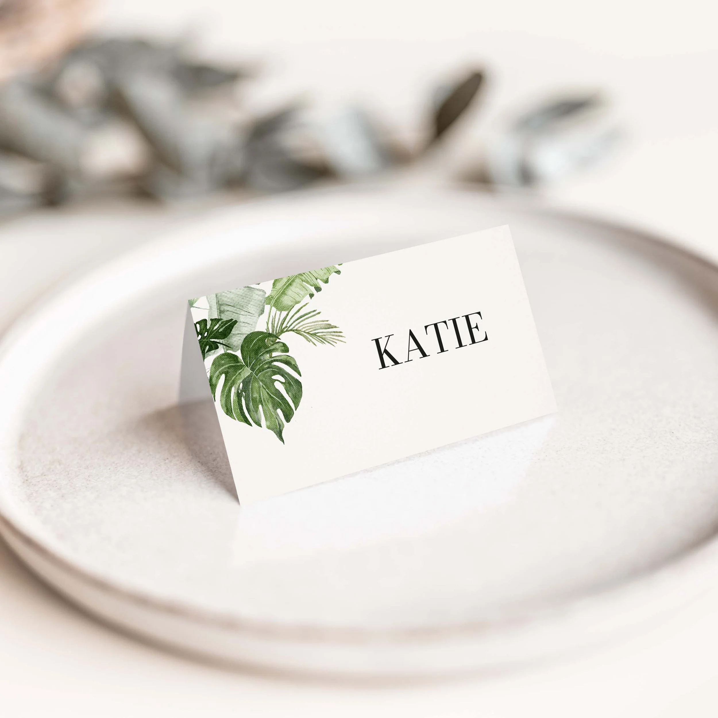 Tropical wedding place card with bold monstera and palm leaf botanical illustration and clean serif guest name typography on luxury off-white card
