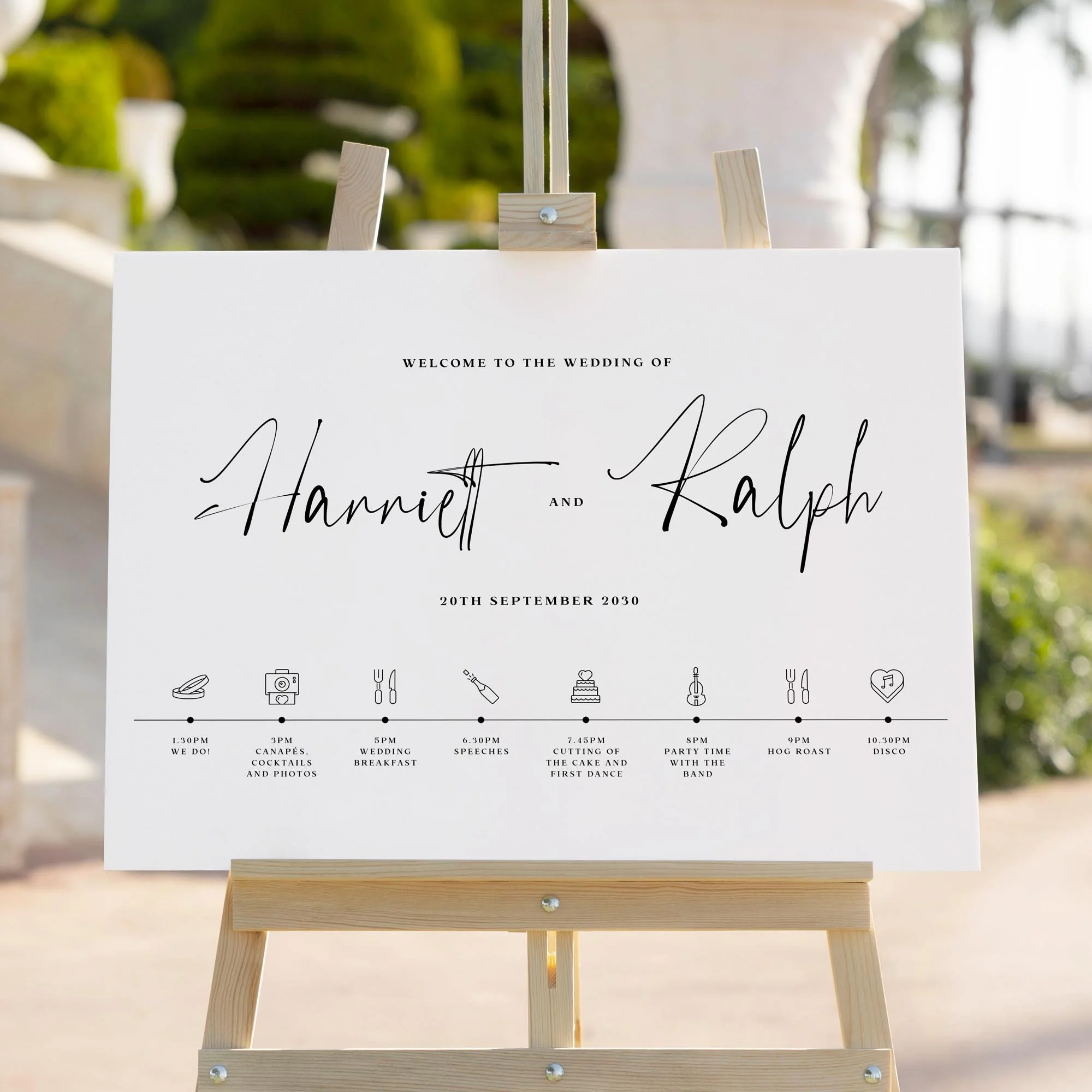 Modern monochrome wedding signage in calligraphy style with order of the day timeline and icons, displayed on a wooden easel with greenery and stone columns in the background.