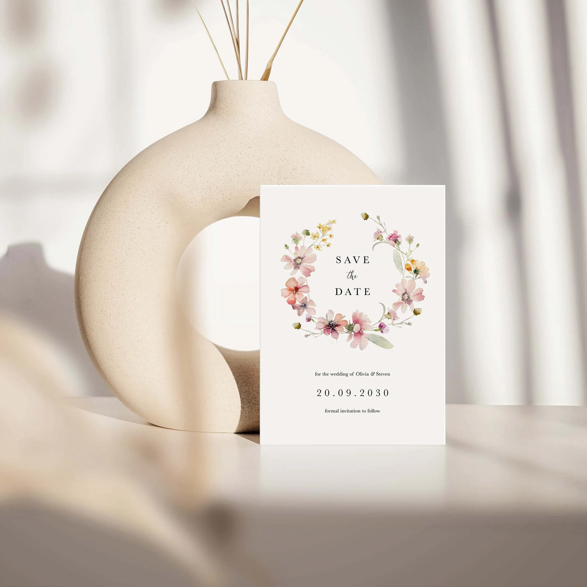 Bloom save the date card with blush pink wildflower wreath design displayed against a cream ceramic vase on a neutral background