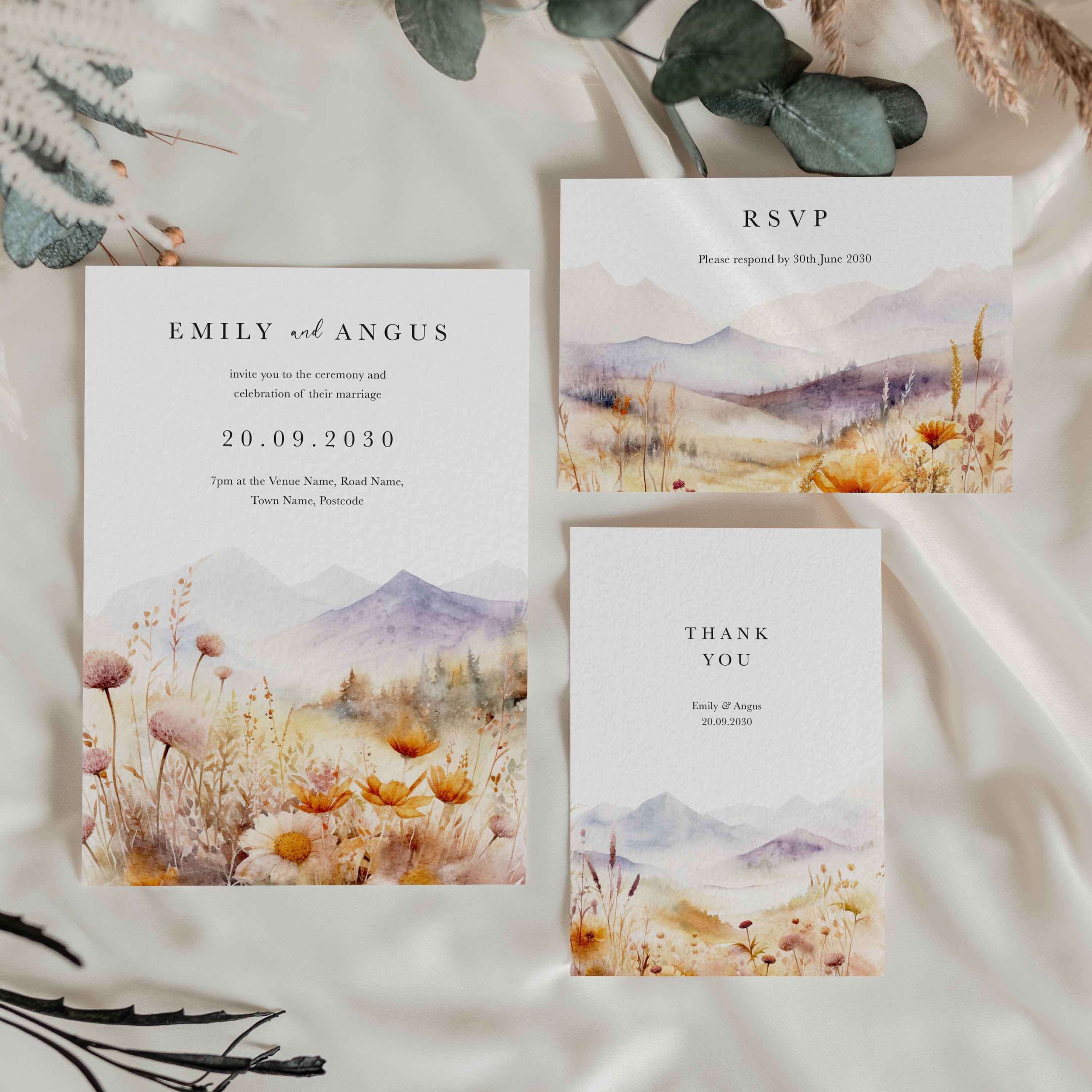 Scenic wedding stationery suite flat lay showing invitations, RSVP cards and coordinating pieces with a soft purple mountain and golden meadow watercolour design