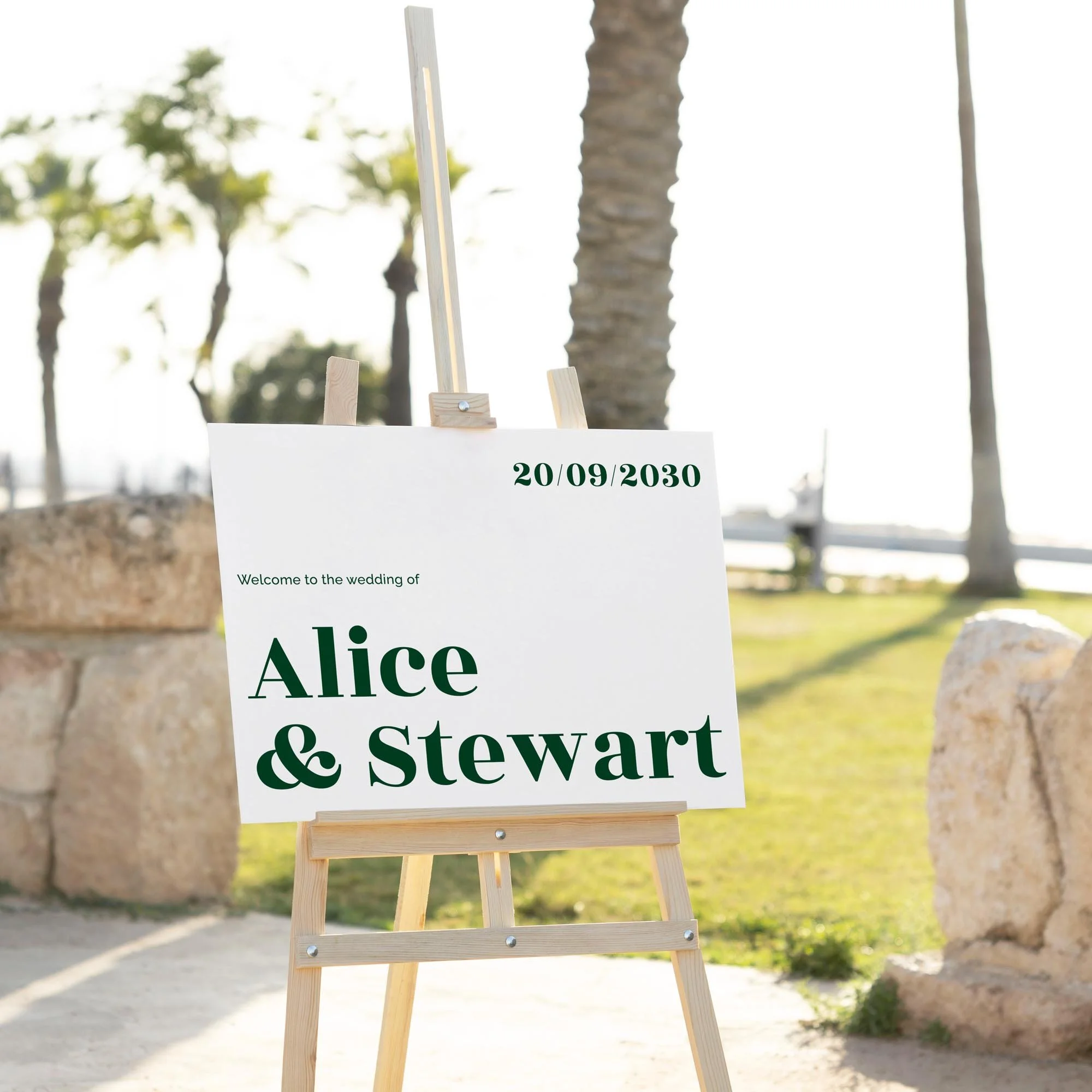 Personalised wedding welcome board featuring bold serif typography in forest green, shown on a wooden easel outdoors with palm trees and stone wall backdrop.