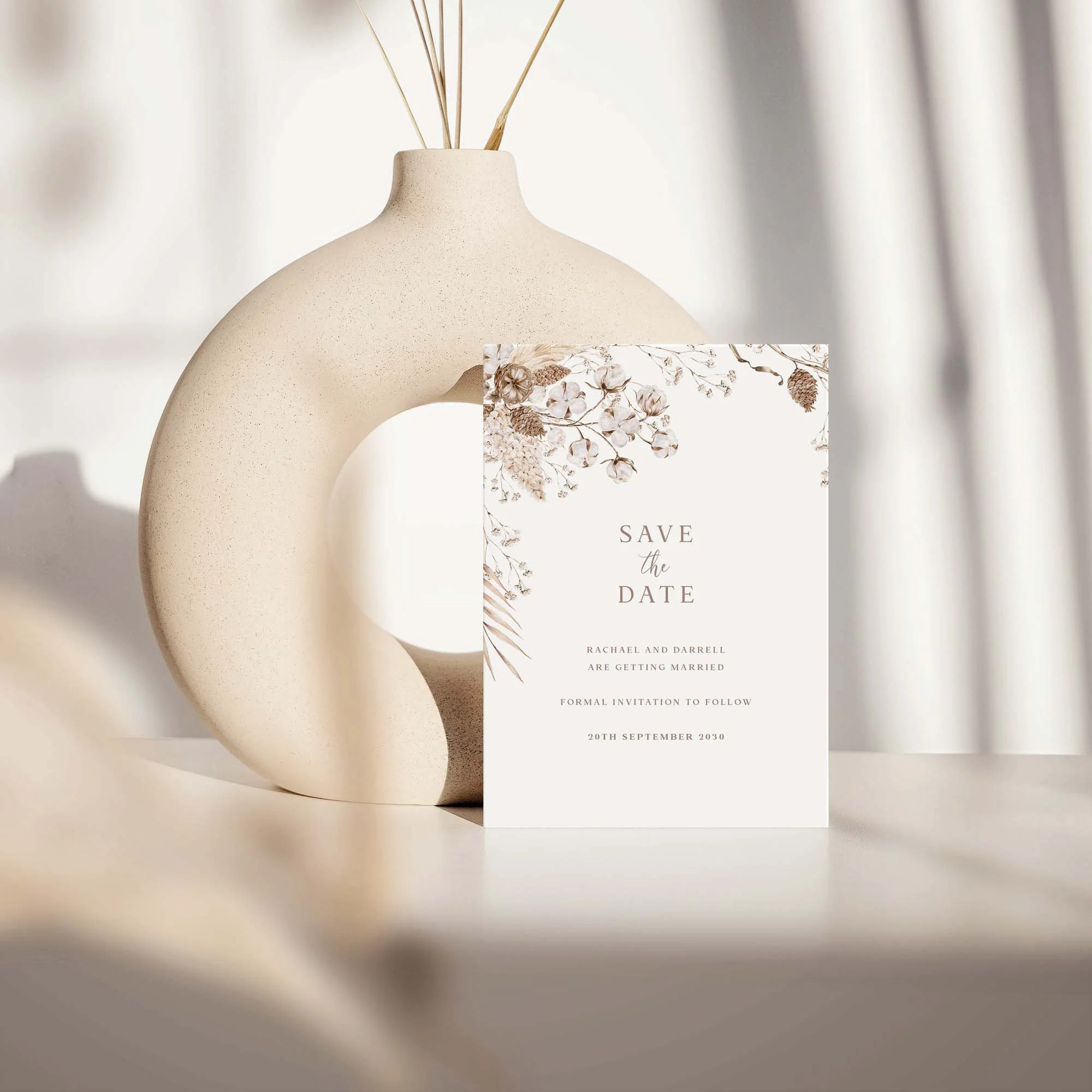Boho save the date card with dried botanical wreath design displayed against a cream ceramic vase on a neutral background
