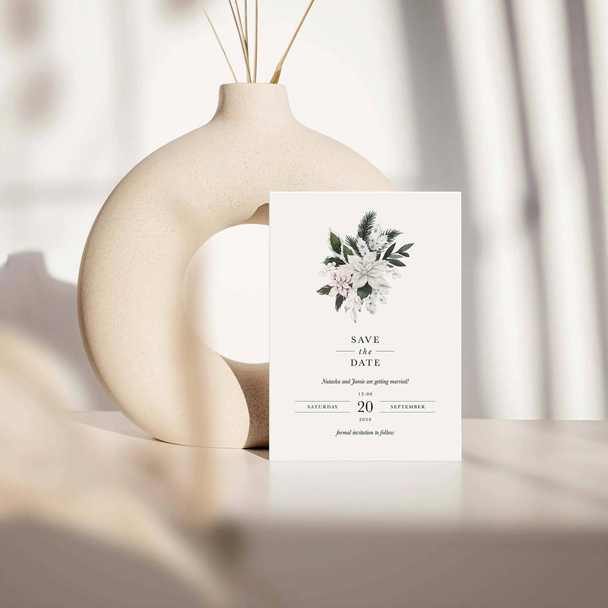 Winter Frost save the date card with white poinsettia botanical wreath displayed against a cream ceramic vase on a neutral background