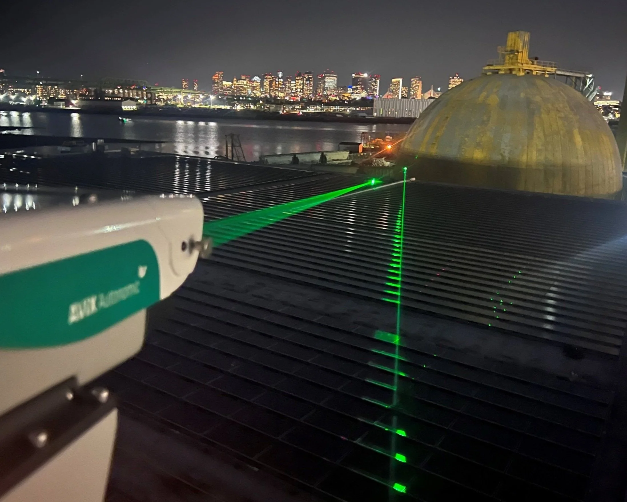 A laser measuring device directing a green laser beam across a rooftop at night, with a city skyline and river in the background.