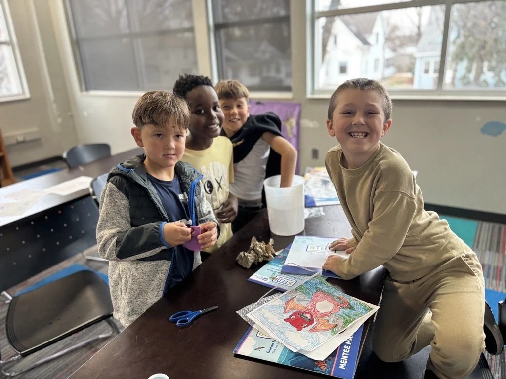 Four children in a classroom around a table, with books and coloring sheets. They are participating in free enrichment after-school programs, smiling and engaged in creative activities and STEM.