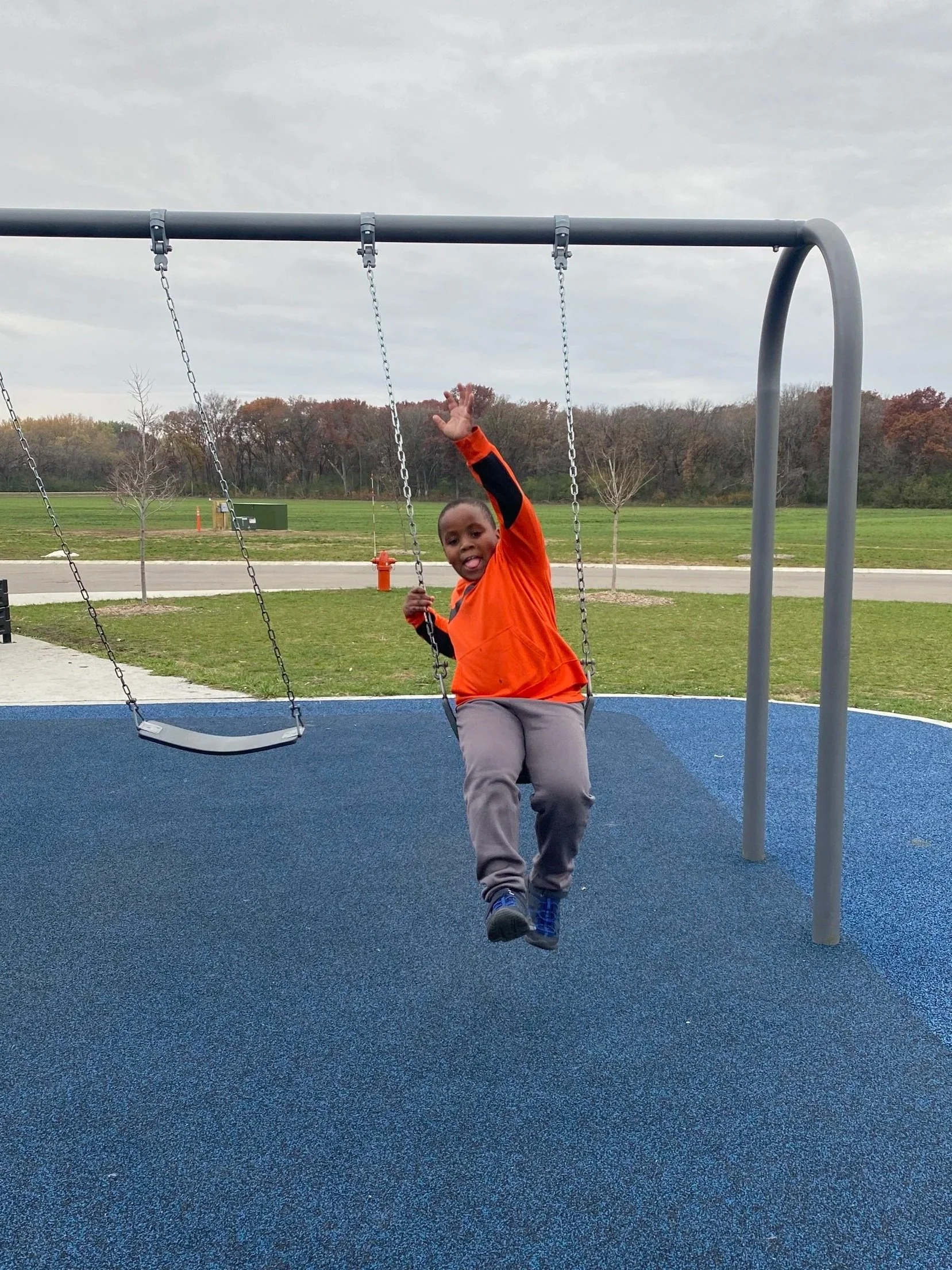 A young boy in an orange shirt and gray pants sitting on a swing at a park, with trees and a cloudy sky in the background.