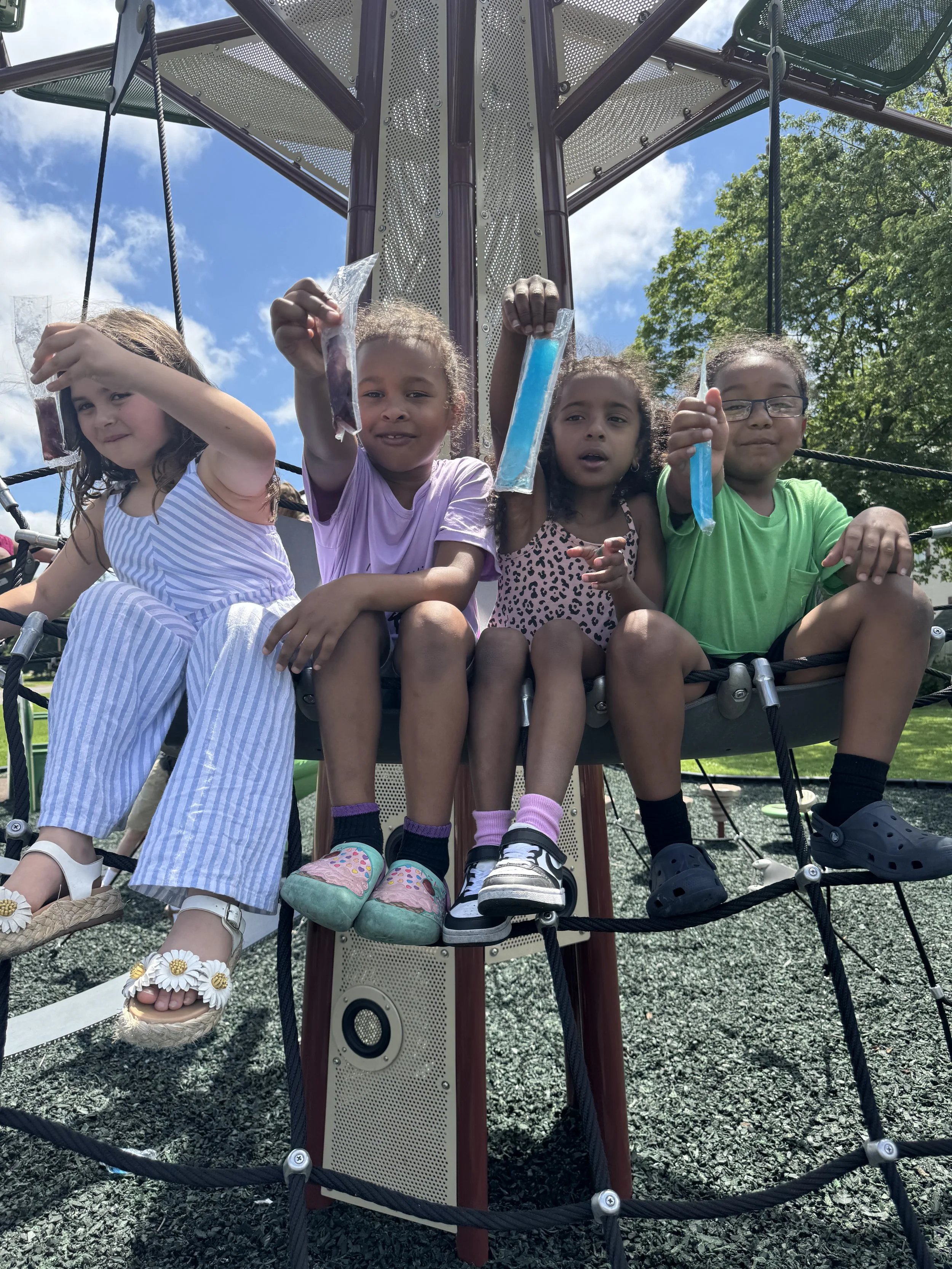 Four young children sitting on a playground structure, holding up colored slime in plastic bags, with a blue sky and green trees in the background.