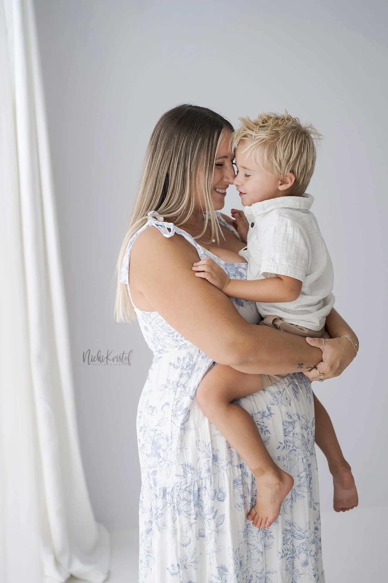 A woman wearing a white floral dress holds a small boy in her arms, both touching foreheads and smiling against a plain gray background.