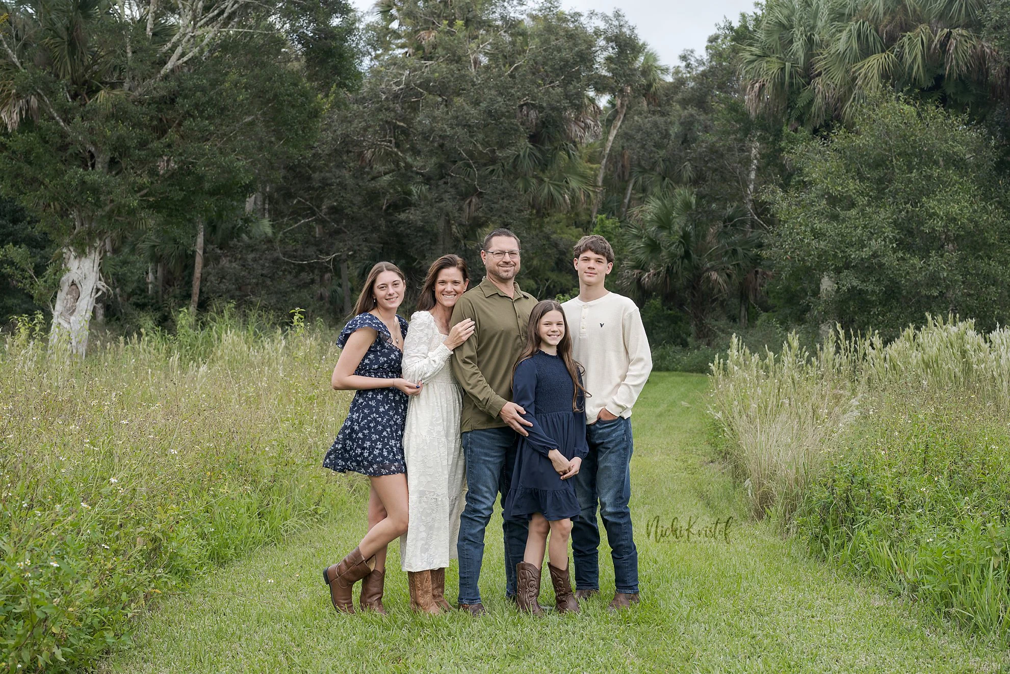 A family of five standing together in a grassy field with trees in the background. The family includes a woman, a man, two girls, and a boy, all smiling and dressed casually.