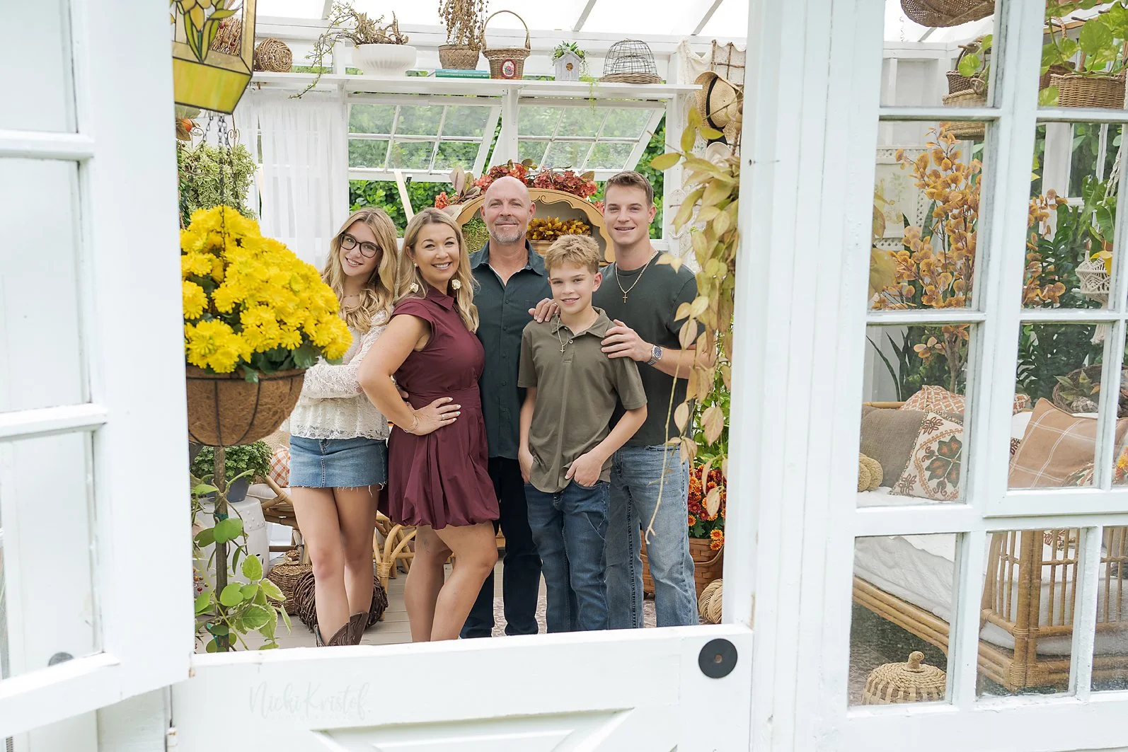 Family of five standing inside a greenhouse, posing for a photo. The group includes a woman in a maroon dress, a man in a black shirt, a teenage boy in a dark t-shirt, and two children, a girl in glasses and a girl in a tan t-shirt. The greenhouse is