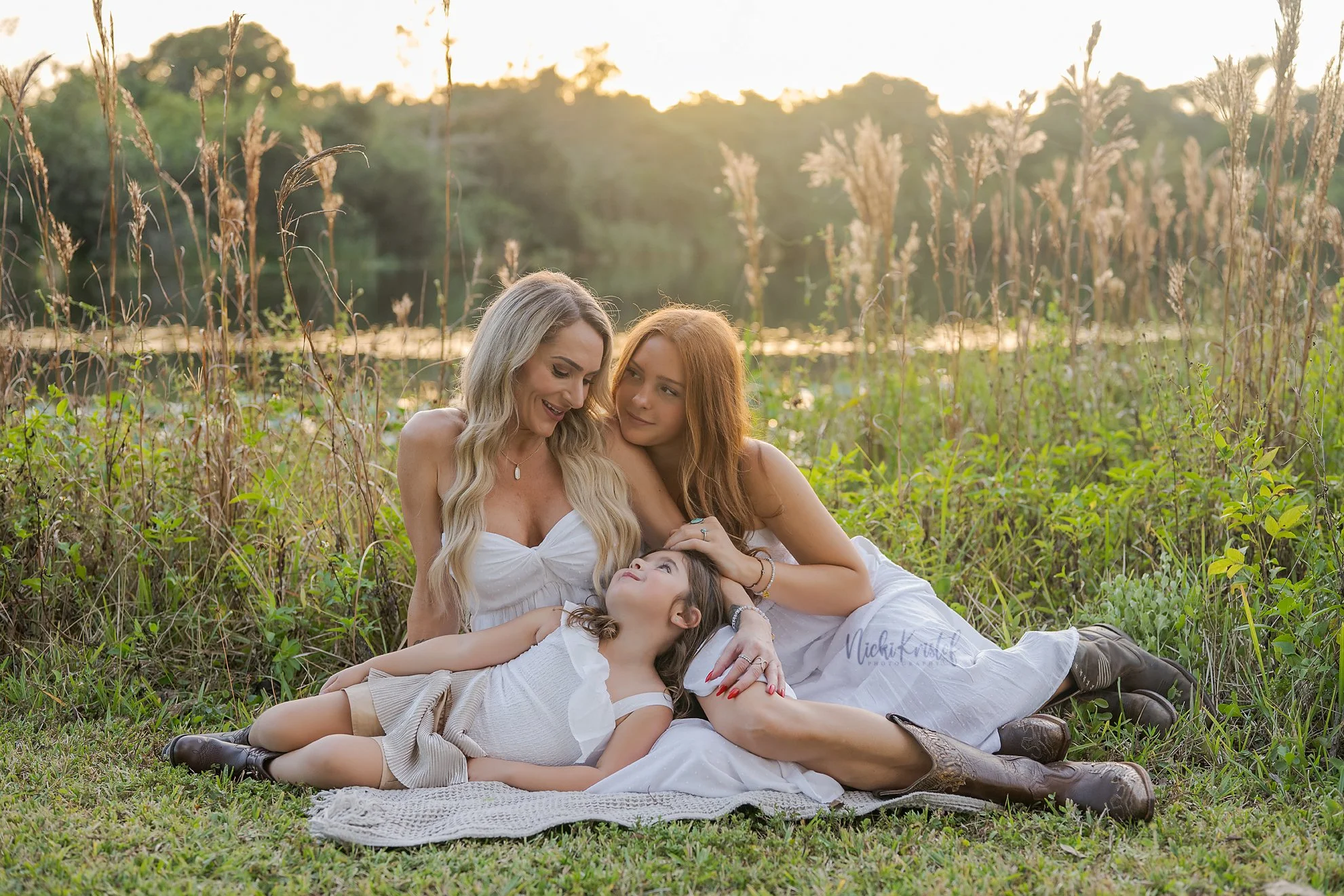A woman with long blonde hair, a red-haired girl, and a young girl with dark hair are lying on the grass by a river at sunset. They are wearing white dresses and appear happy and relaxed in nature.