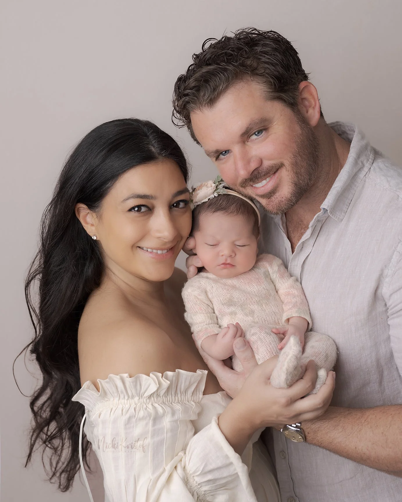A happy mixed-race family of three, including a woman with long dark hair, a man with curly brown hair and light skin, and a newborn baby girl dressed in a pink outfit with a floral headband. The family is smiling and embracing each other against a p