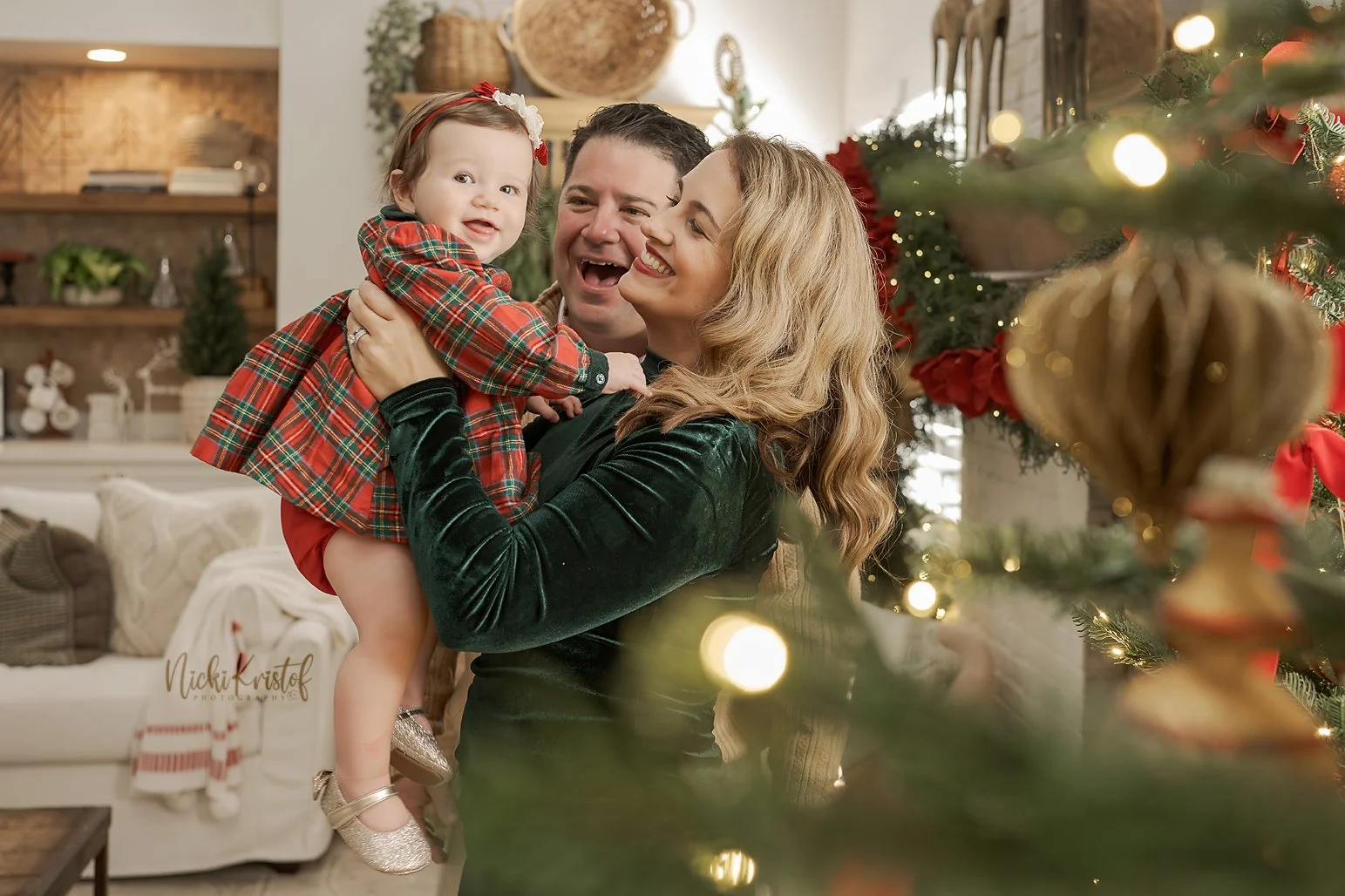 A happy family of three celebrating Christmas indoors, with a decorated Christmas tree and holiday decorations in the background. The woman is holding a smiling young girl dressed in a red plaid dress.