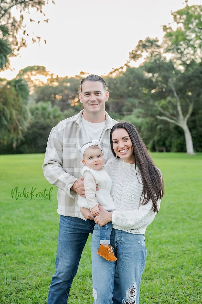 A happy family of three outdoors in a park during sunset, with a man, woman, and a young girl holding a toddler.