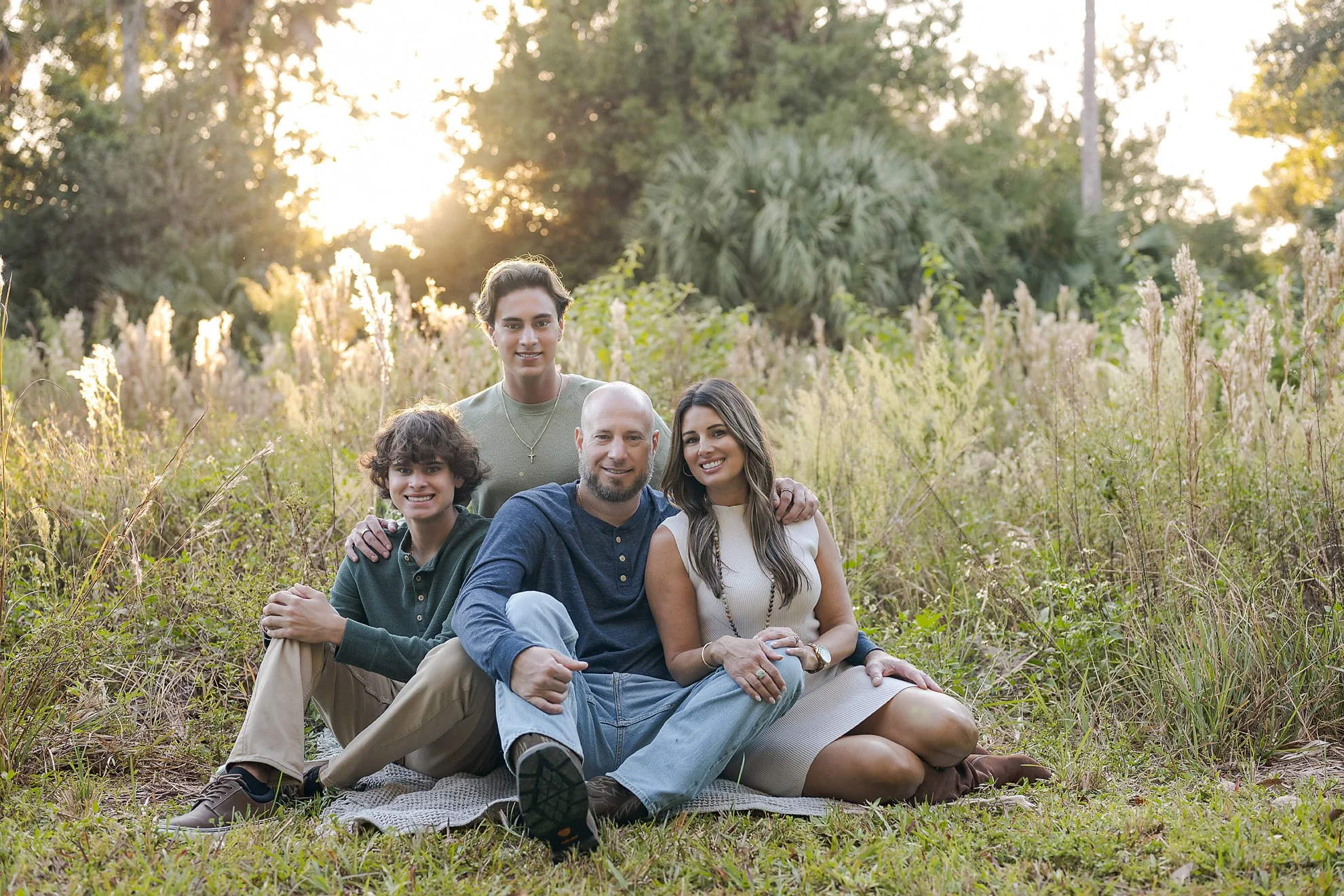 A family of five sitting on a blanket in a grassy field with trees and sunlight in the background, enjoying the outdoors during sunset.