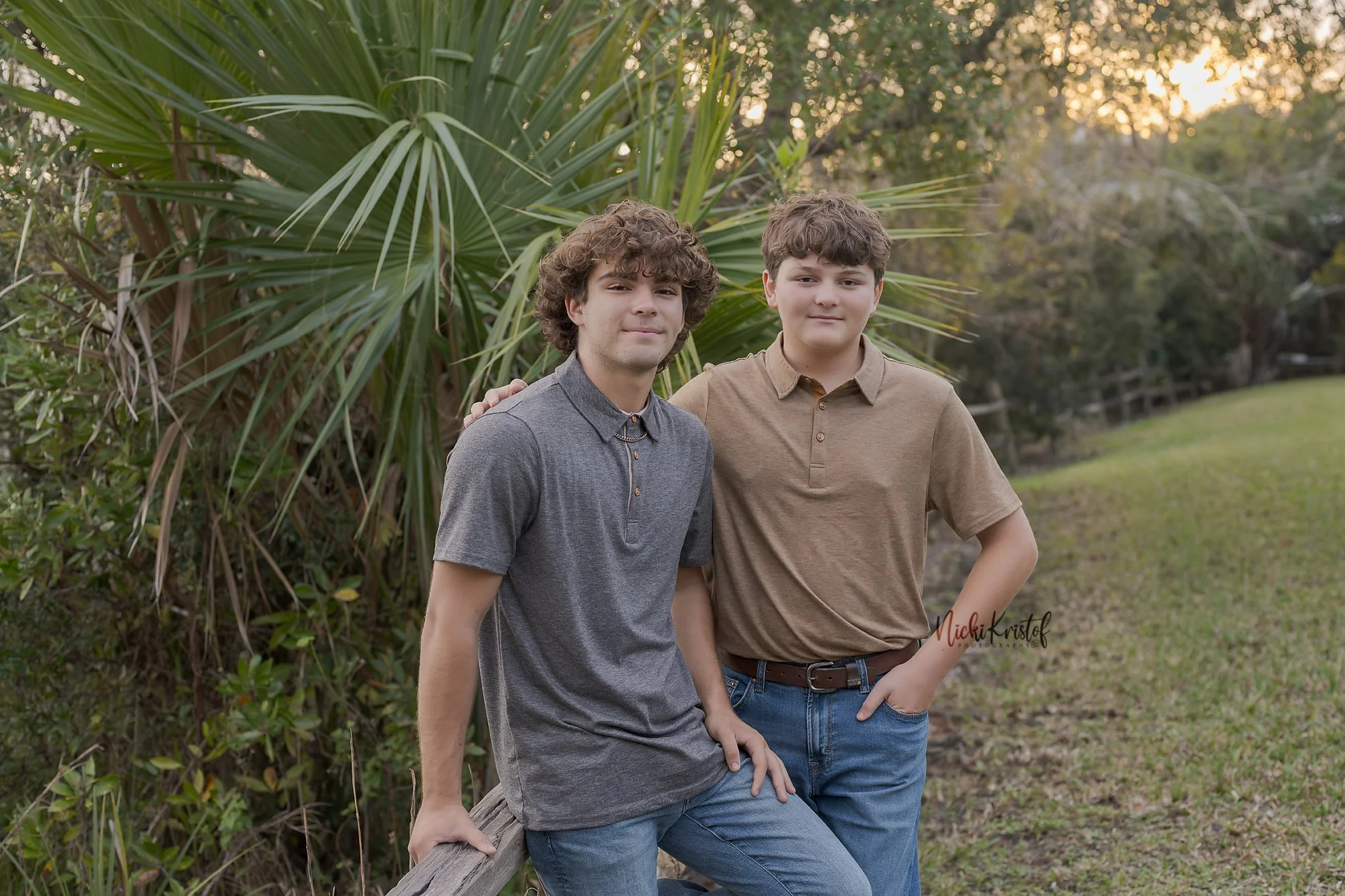 Two young boys with curly hair standing outdoors near palm plants and trees during sunset, smiling at the camera.