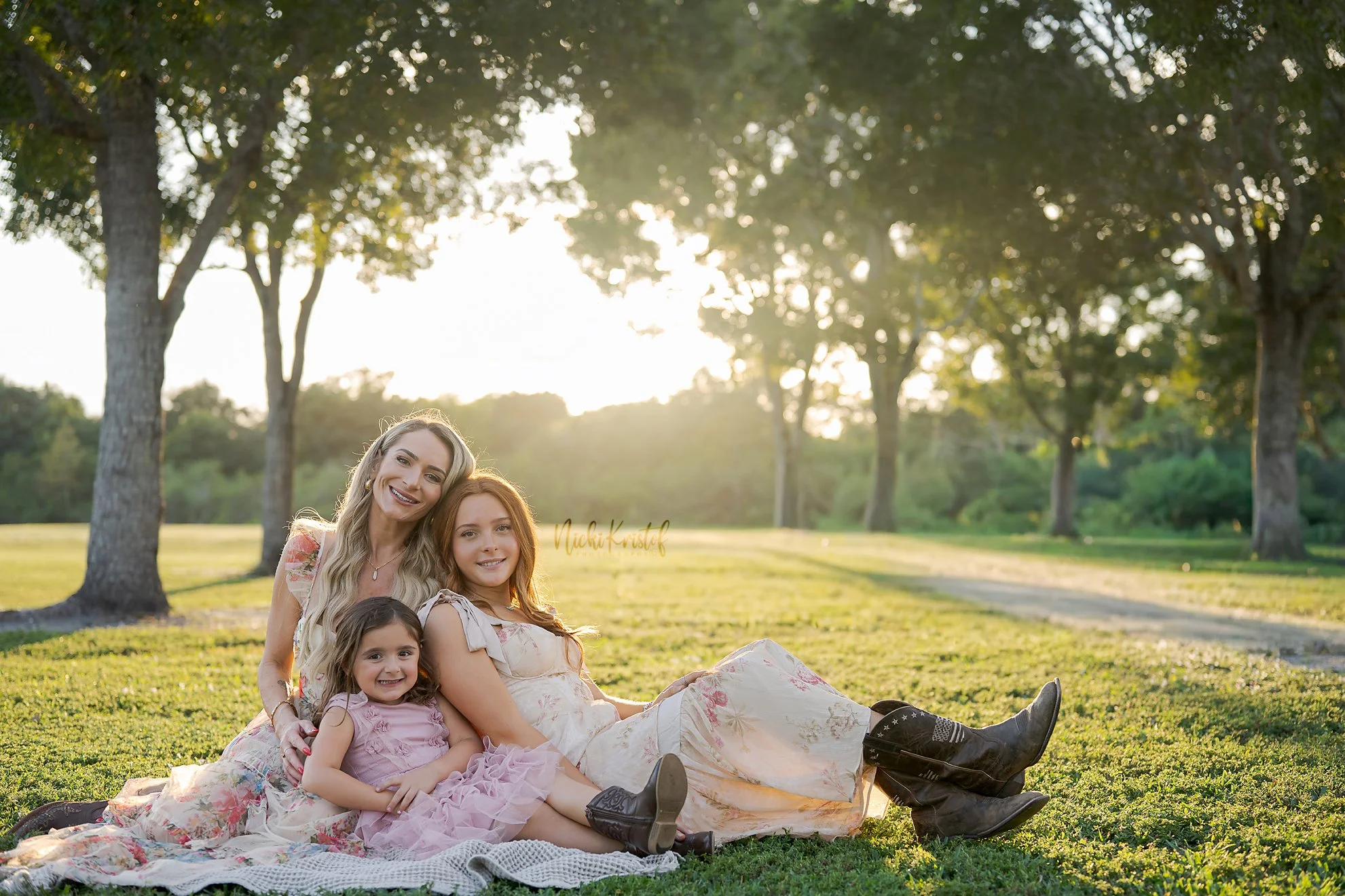 A group of five women and girls sitting on a blanket in a park, smiling in the sunlight with trees in the background.