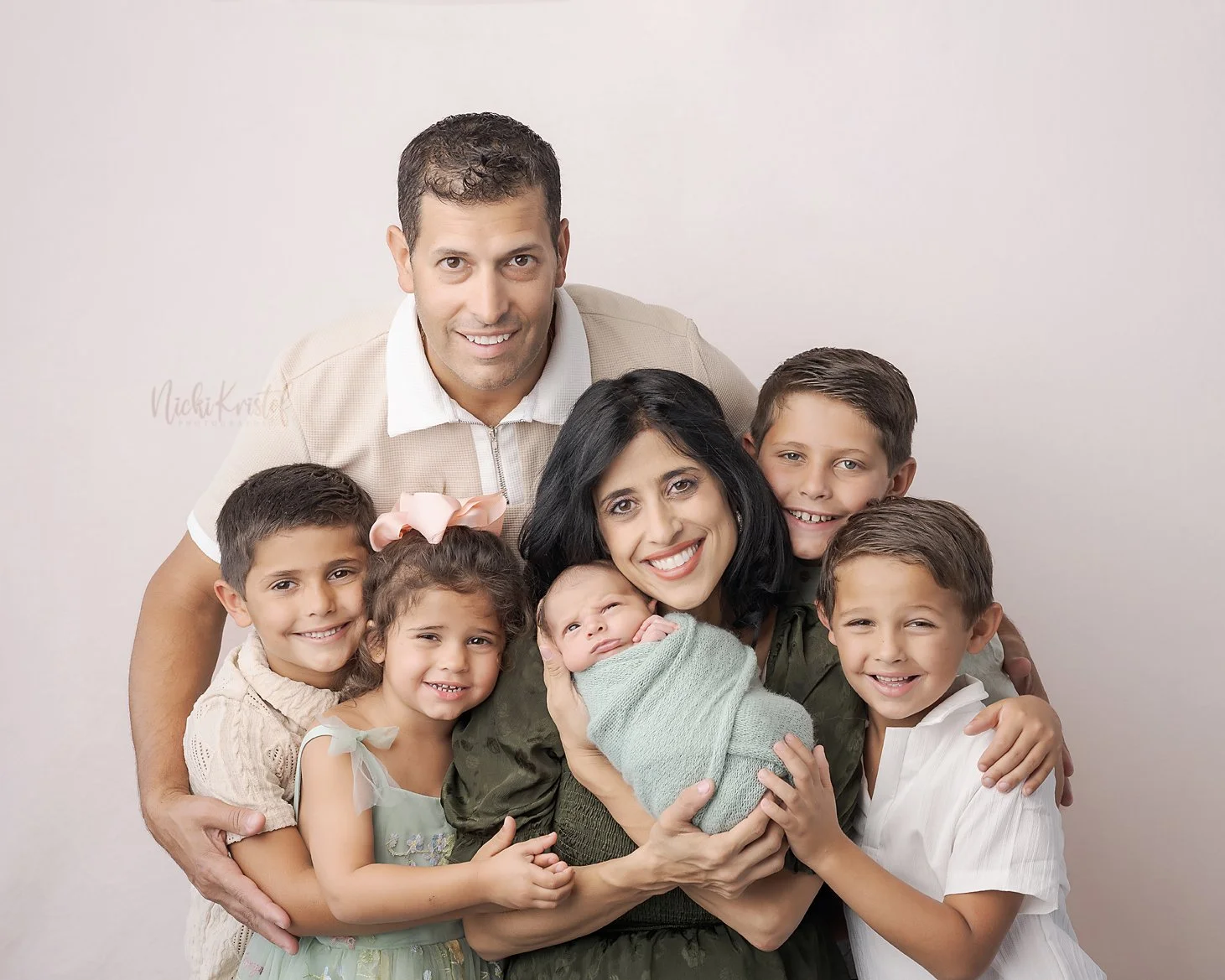 A family of eight, including a father, mother, and six children, all smiling and hugging, with the mother holding a newborn wrapped in a blanket.