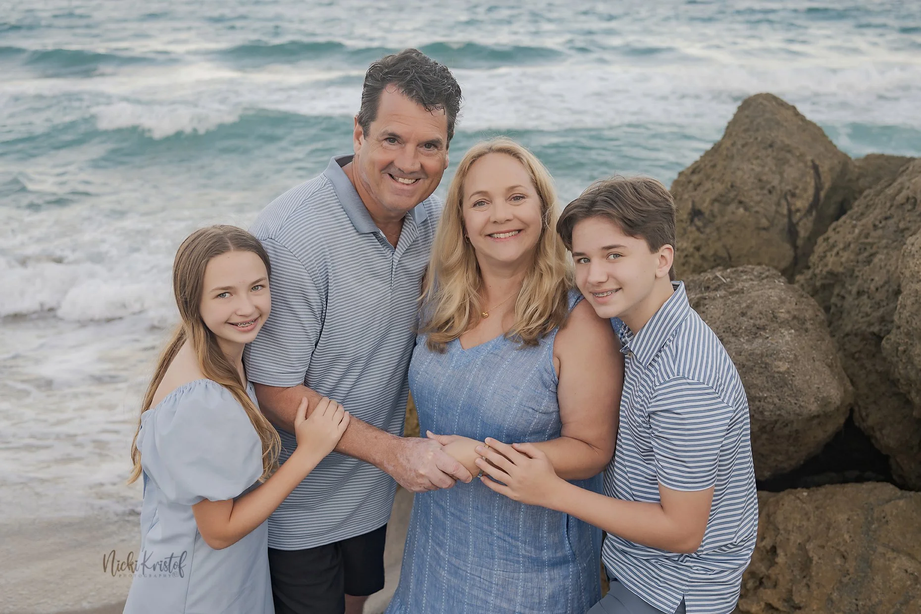 A family of five stands together on a beach by the ocean with rocks in the background. They are smiling and holding hands, wearing casual summer clothing.