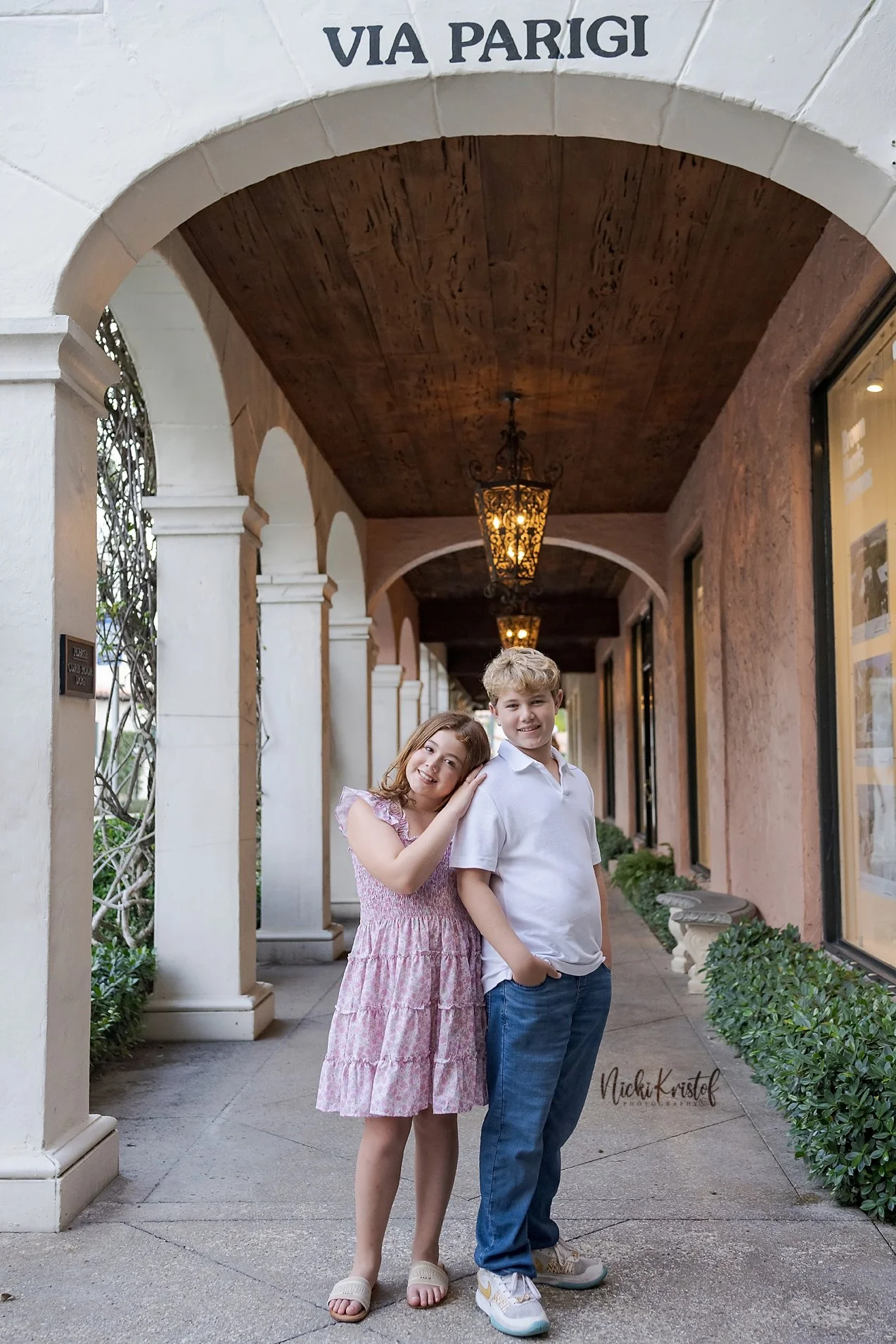A young girl and boy standing together in a covered walkway with columns, hanging lanterns, and greenery, smiling at the camera.