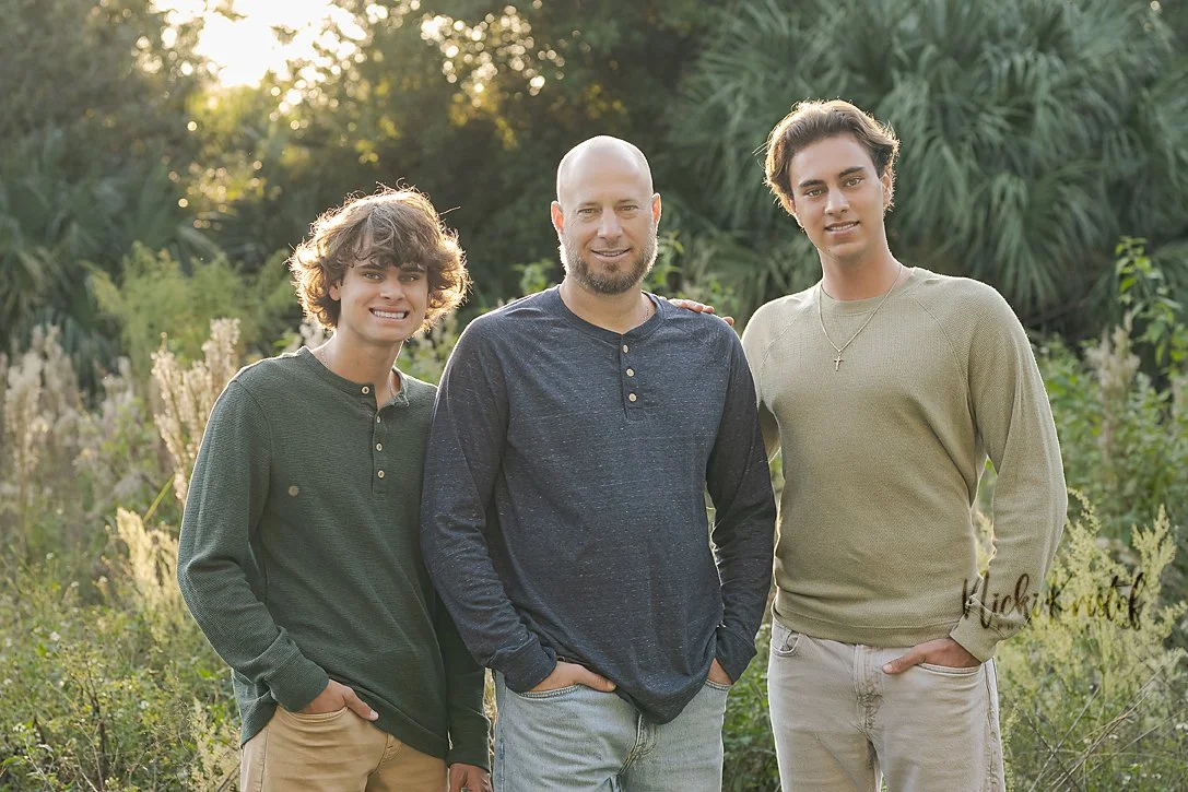 Three men standing outdoors in a park or garden with green foliage, smiling at the camera during the daytime.