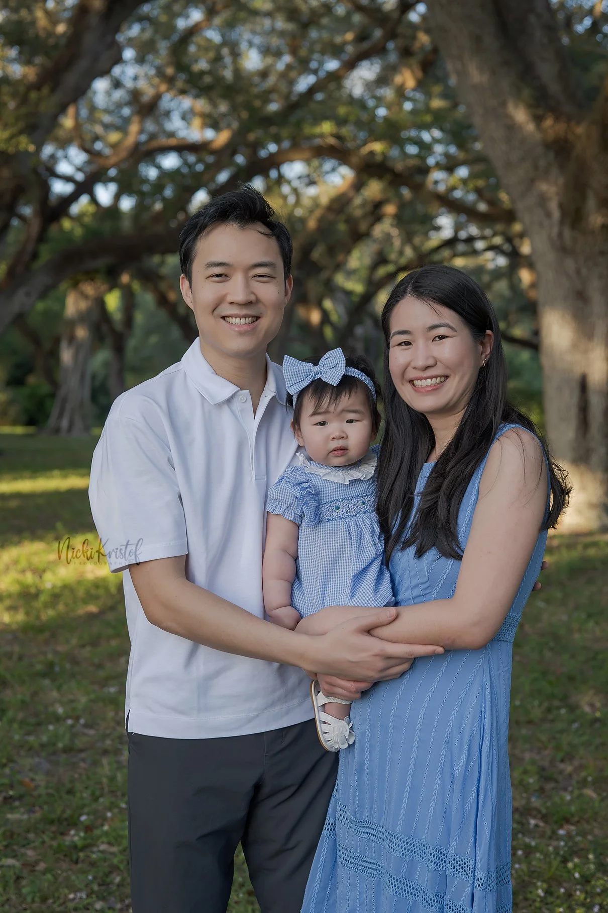 A happy family of three standing outdoors in front of large trees, with the father on the left, the mother on the right holding a young girl, all smiling at the camera.