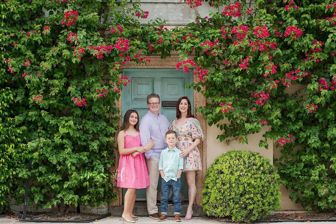 Family of four standing in front of a blue door surrounded by pink flowering bushes and green foliage.