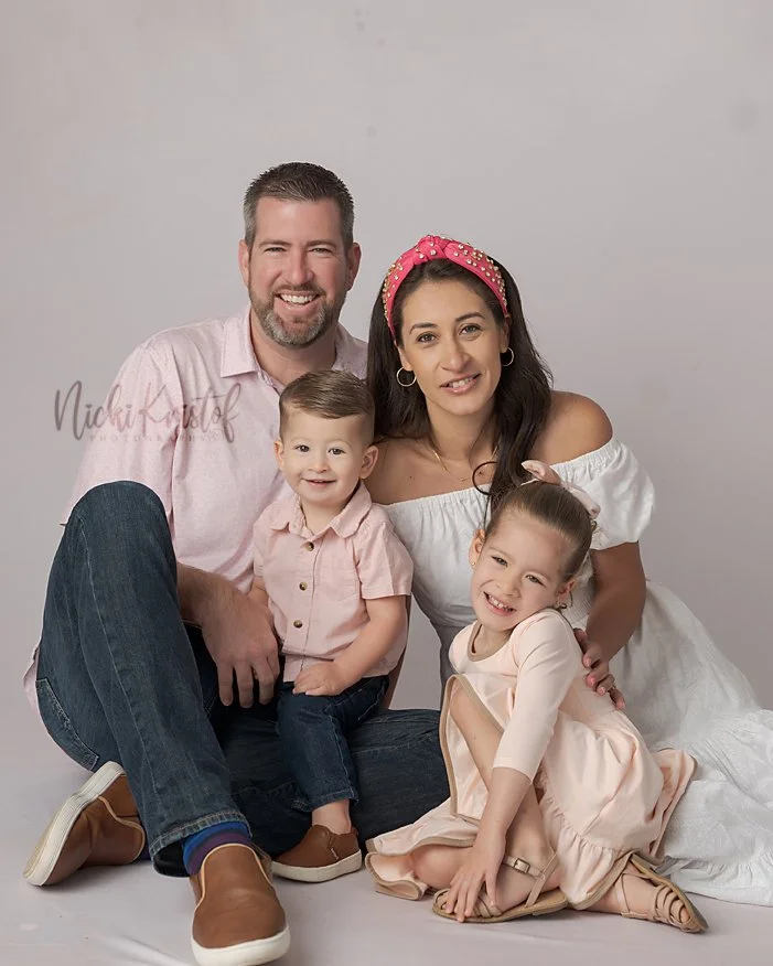A family of four sitting together for a portrait, smiling at the camera. The father has short hair and a beard, wearing a light pink shirt and jeans. The mother has long dark hair, wearing a white dress and a pink headband. The son is sitting on the 